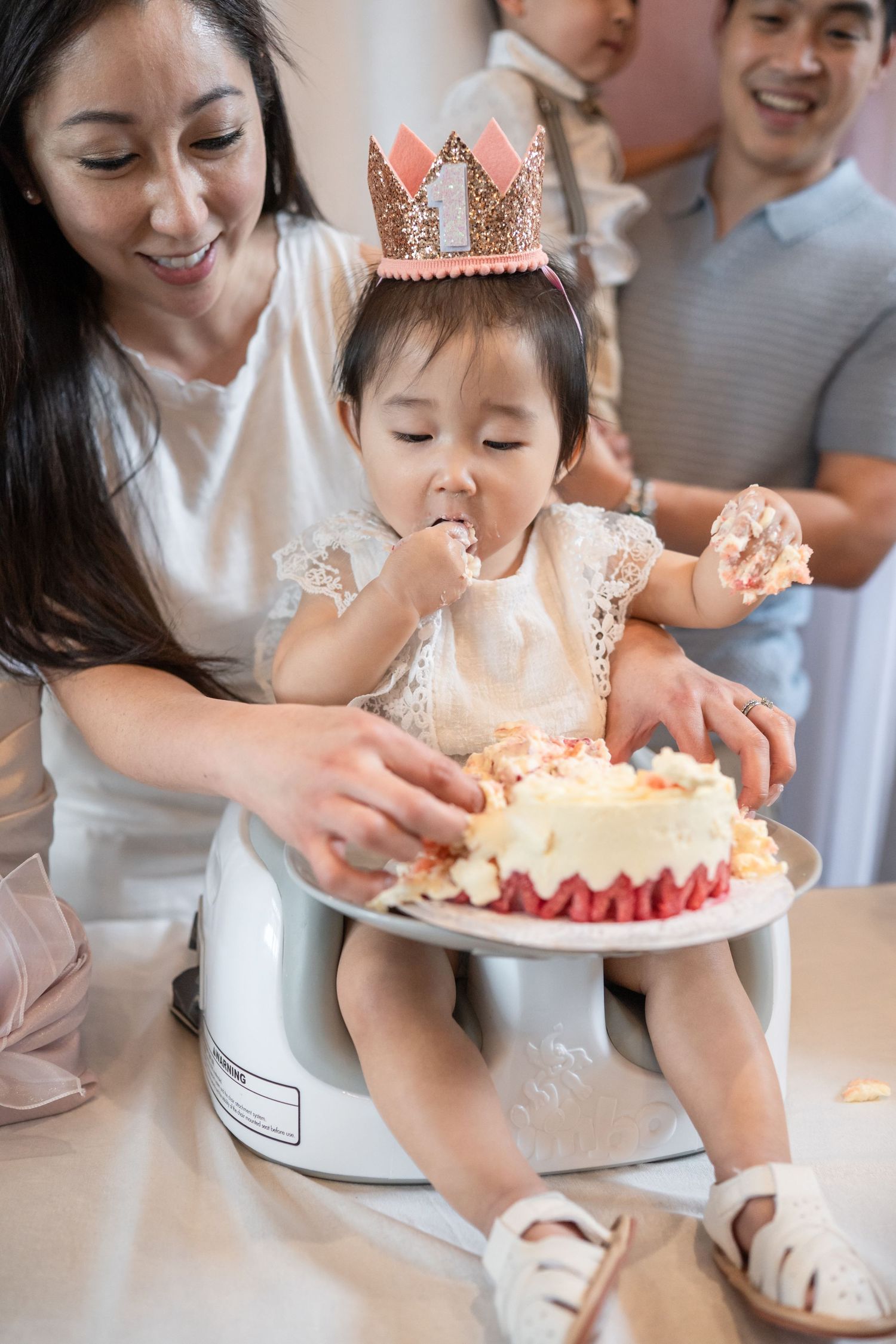Family celebrating with a birthday cake decorated with pink crown and candles on a white cake stand. Dohl Cake smash photographer Reston