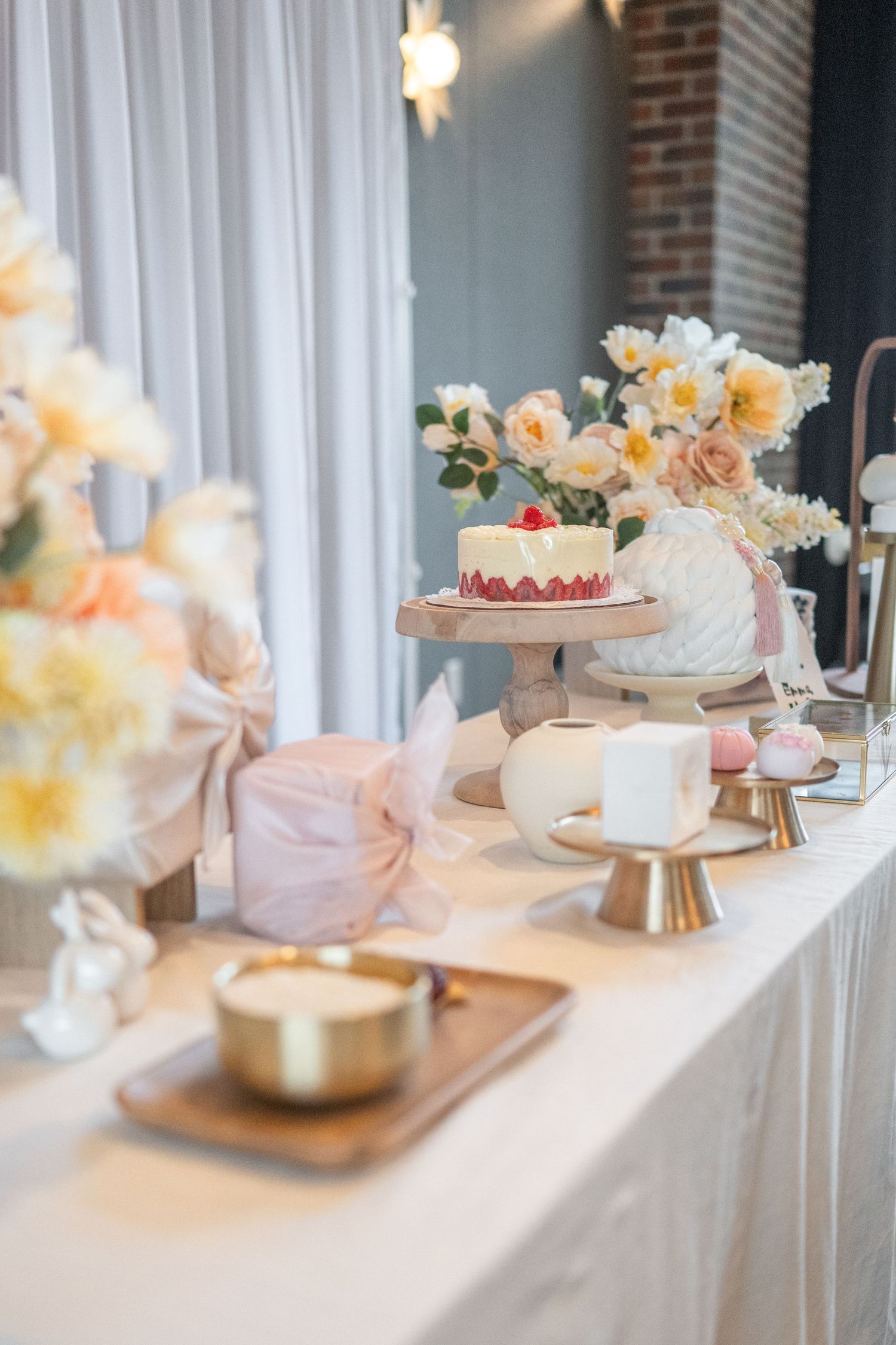 An elegant display of white dohl cakes adorned with delicate flowers set on a white tablecloth against a brick wall. Herndon photographer