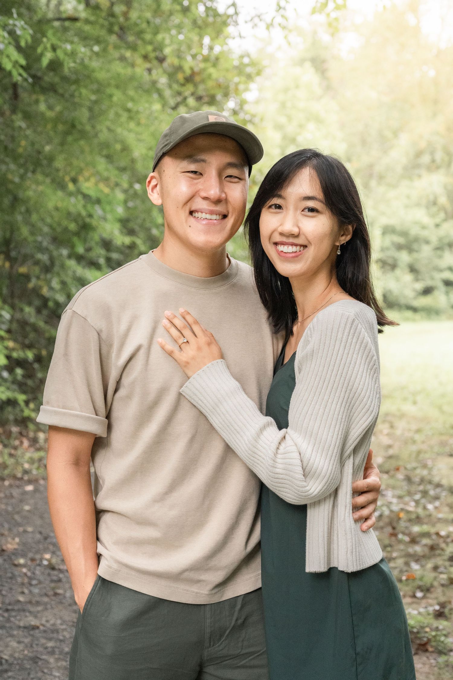 A couple wearing coordinated beige and green outfits embrace and smile while posing outdoors in a wooded setting.
