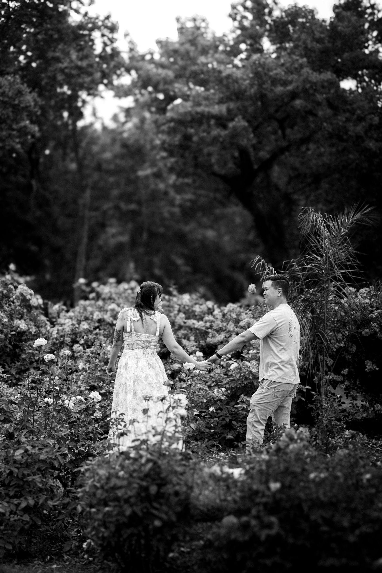 A romantic couple holds hands while standing in a lush garden with blooming flowers in black and white. NoVA Family Photographer