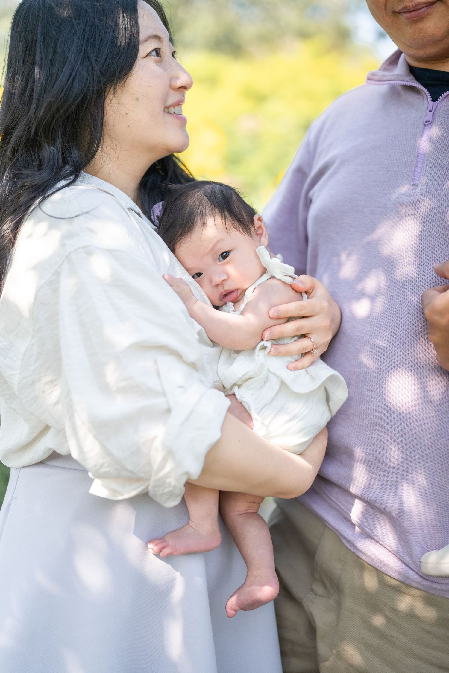 A family shares tender moments while holding their baby in a sunlit outdoor setting wearing white and neutral tones. Alexandria photographer