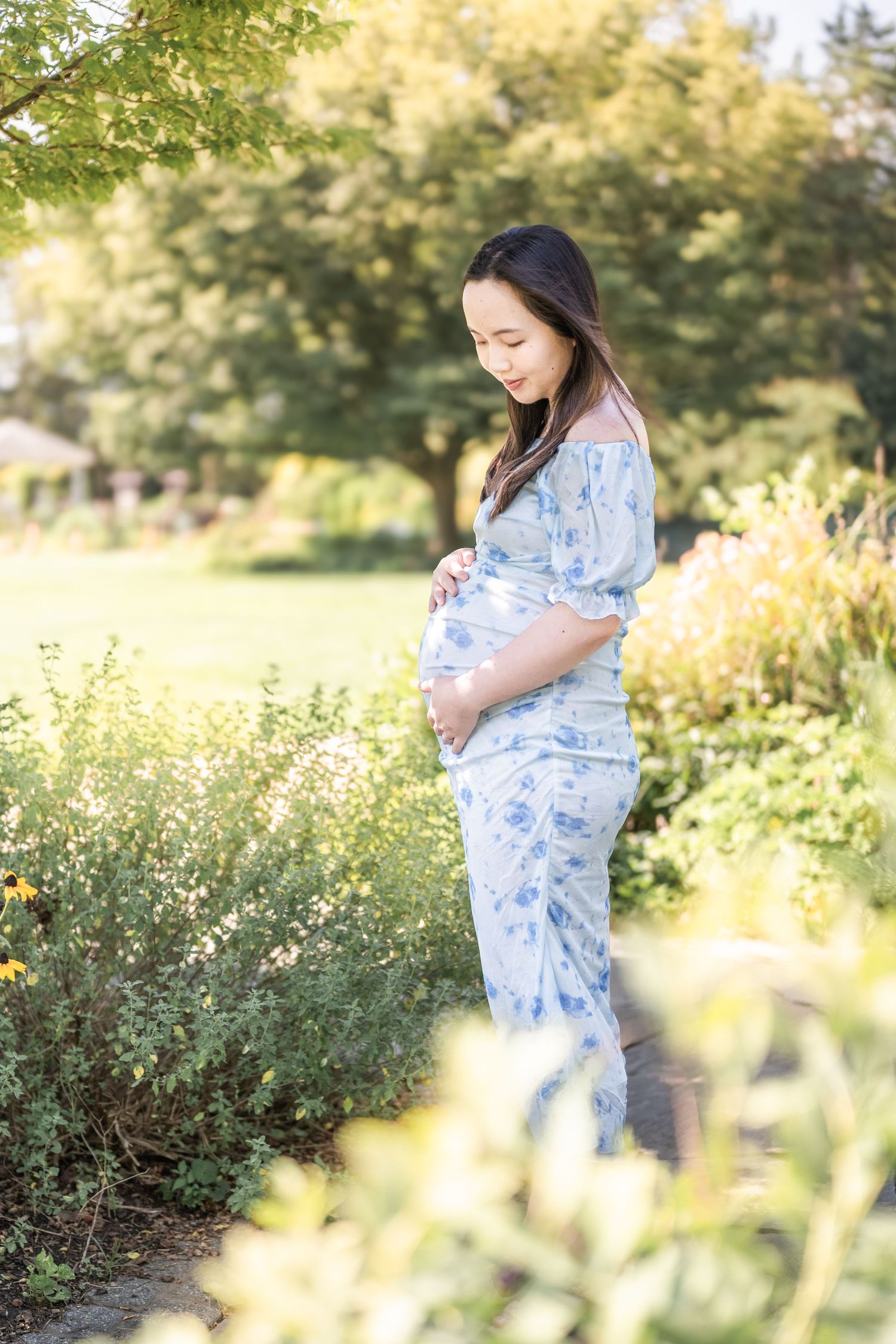 A beautiful maternity photo captures someone in a blue floral dress standing in a sunny garden surrounded by greenery. Reston photographer