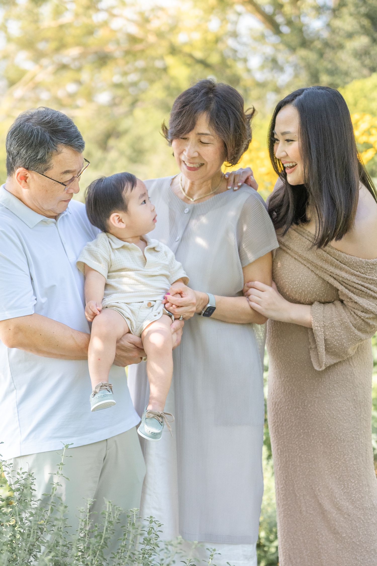 A laughing family in light neutral colors stands together during an outdoor photo session in golden sunlight.