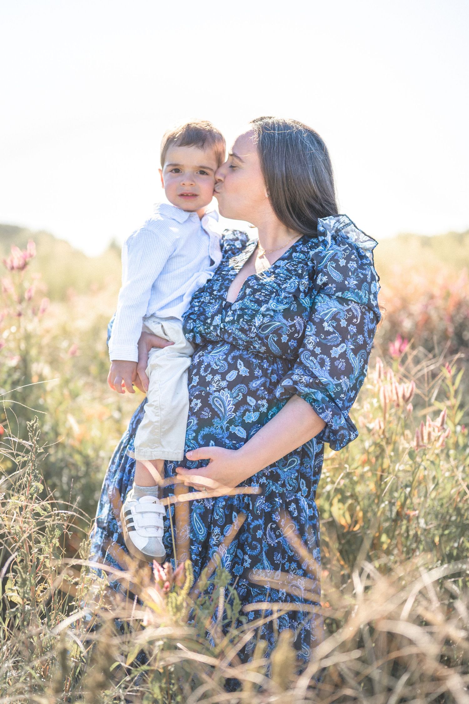 Woman in blue floral dress holding small child in white shirt amidst golden field at sunset.