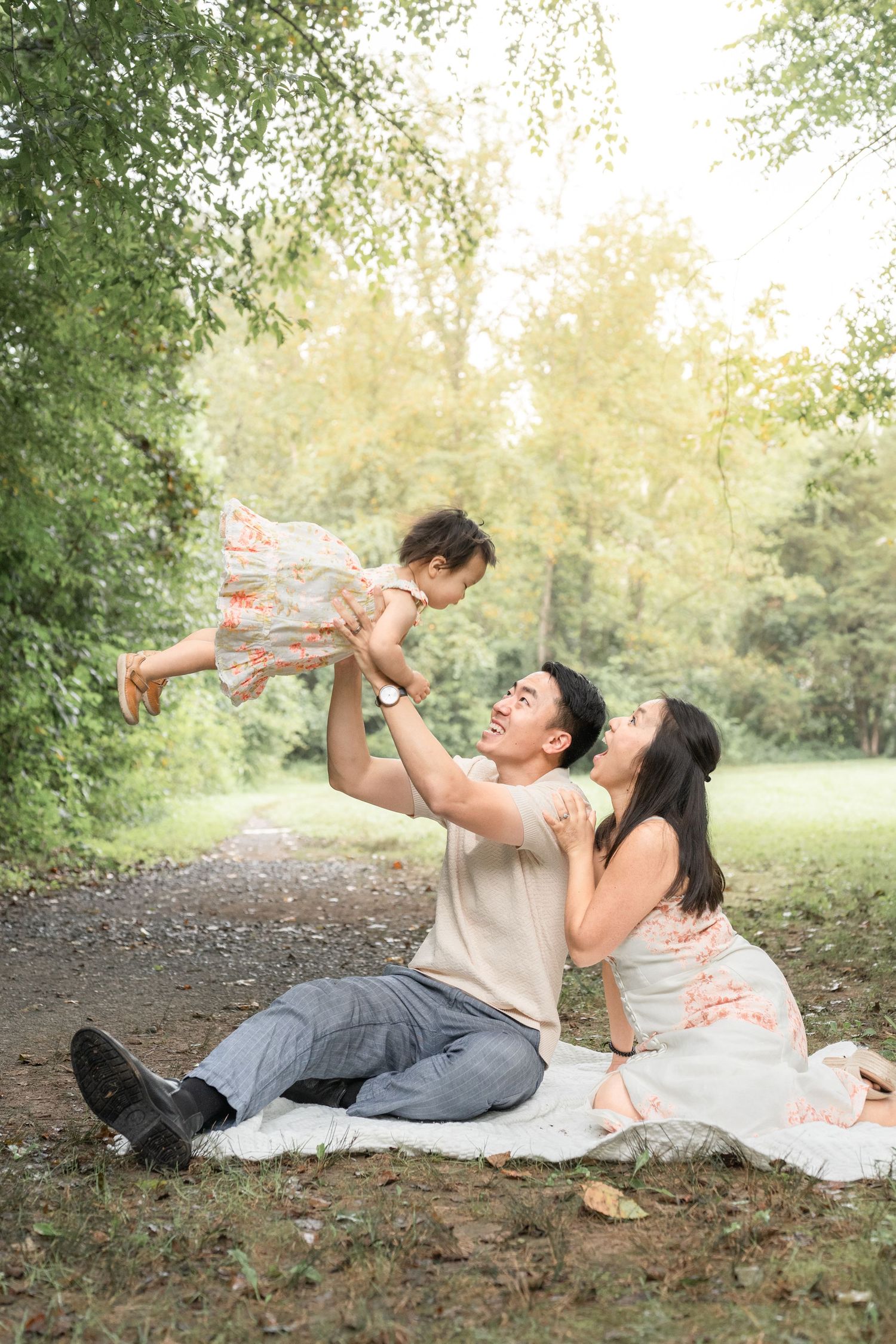 A couple lying on a picnic blanket in a park setting playfully lifting their baby in the air together.