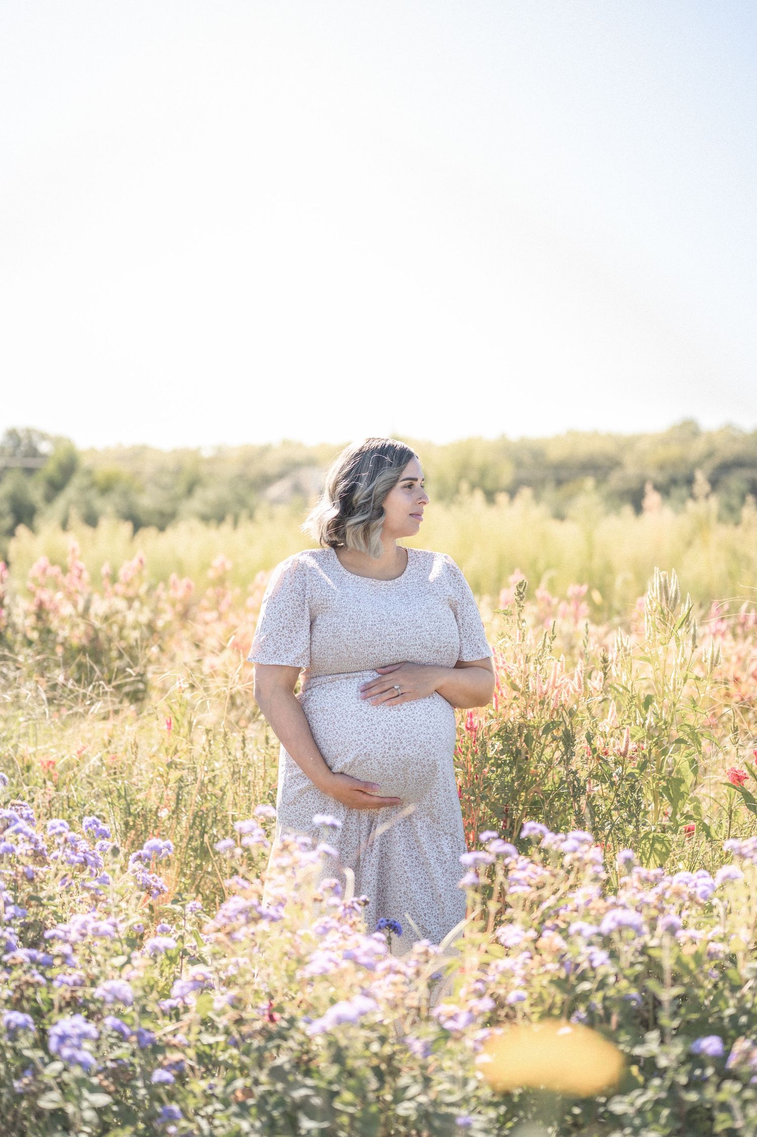 Expecting mother in a white dress tenderly holds her belly while standing in a sunlit meadow with wildflowers.