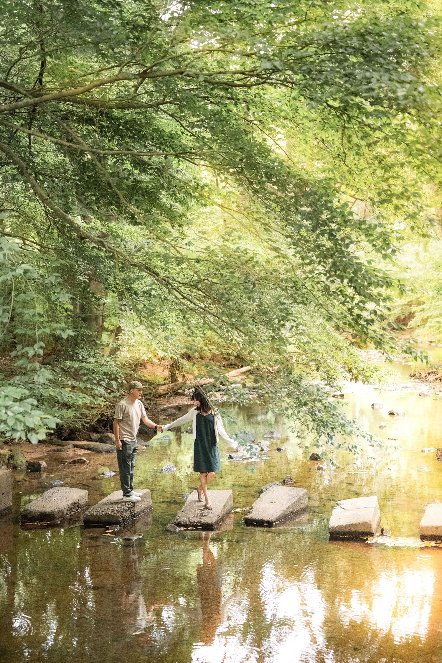 Two people stand on stepping stones in a tranquil forest stream with overhanging trees creating a serene atmosphere.