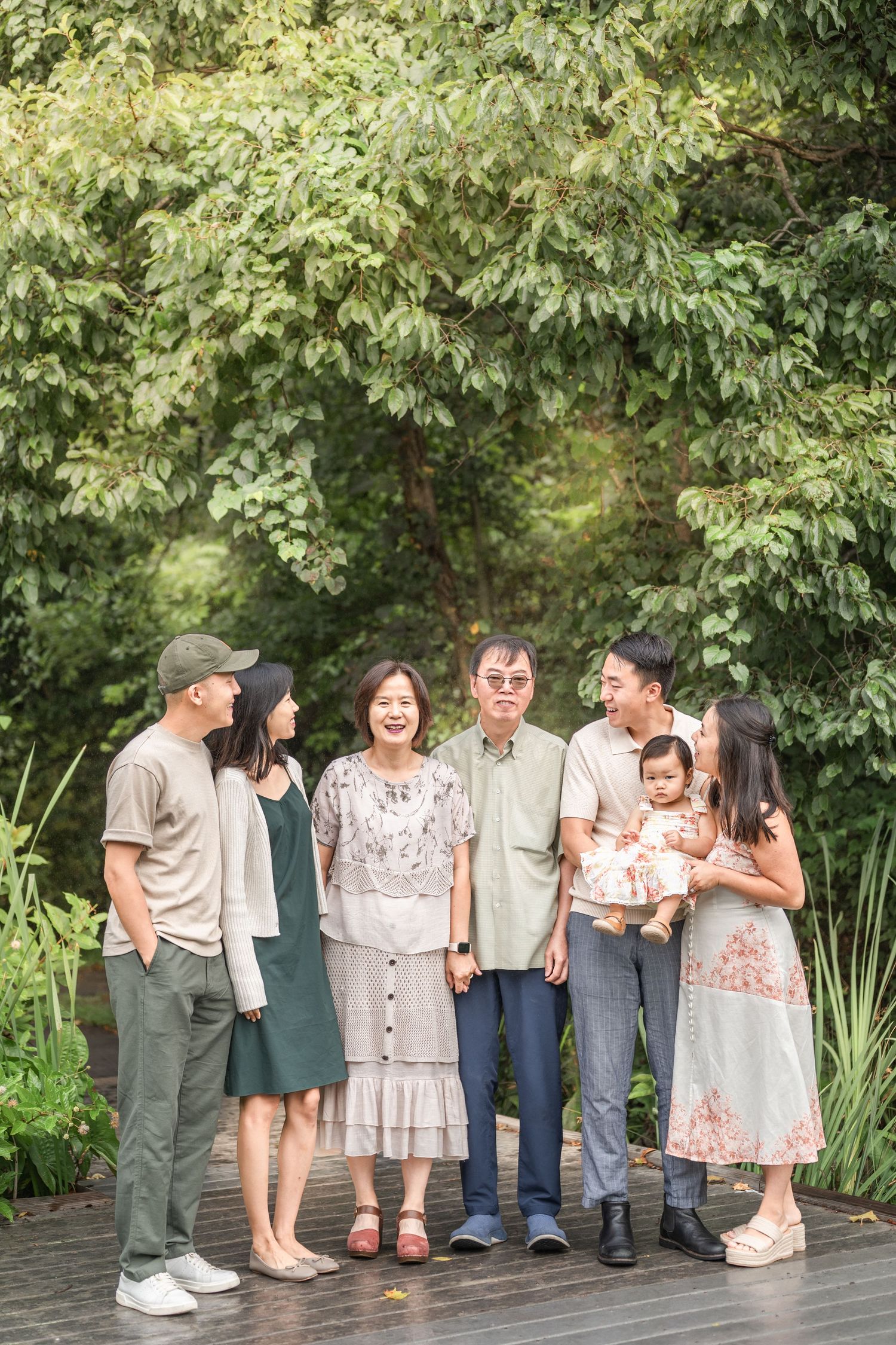 A group of people stand together on a wooden deck in front of green foliage wearing casual clothing.