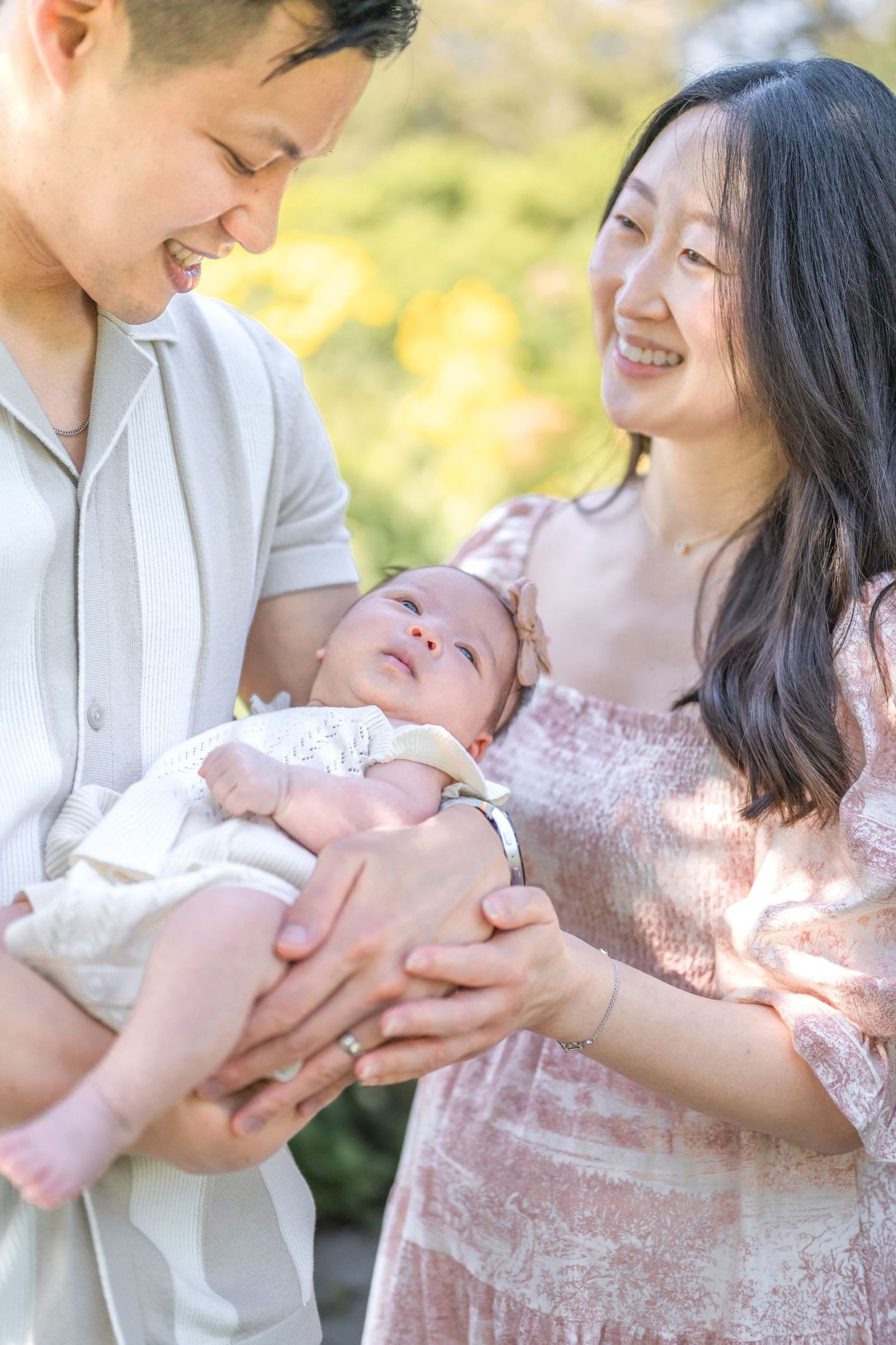 A couple gazes lovingly at their newborn baby outdoors on a sunny day. Photographer