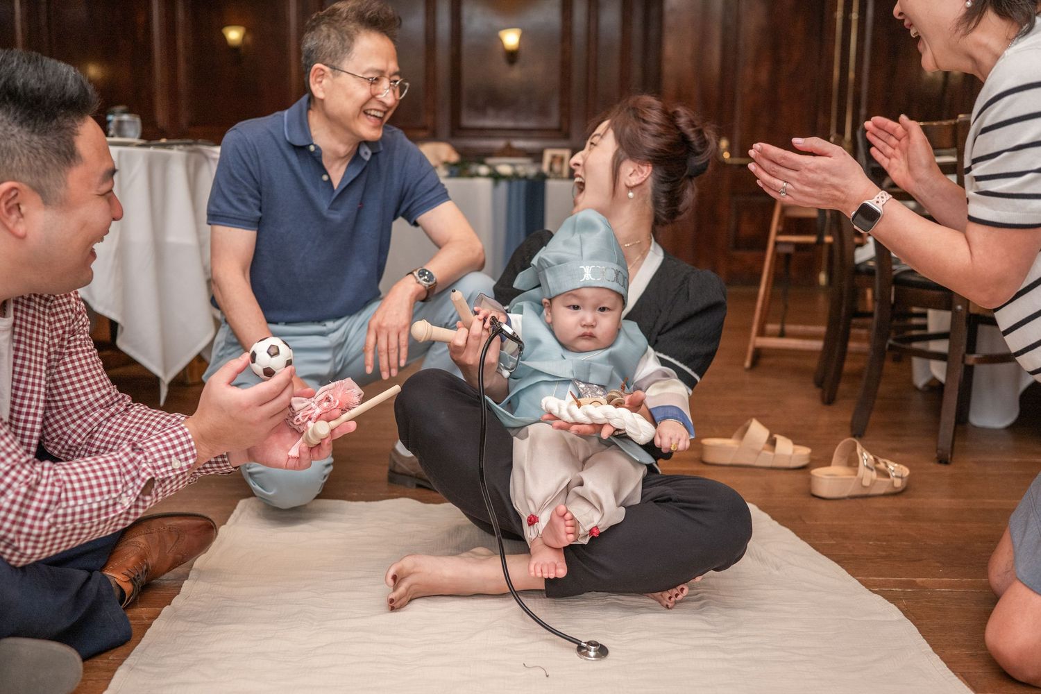 Several people gathered indoors playing a game with cards on a wooden table at a casual social gathering. Dohl
