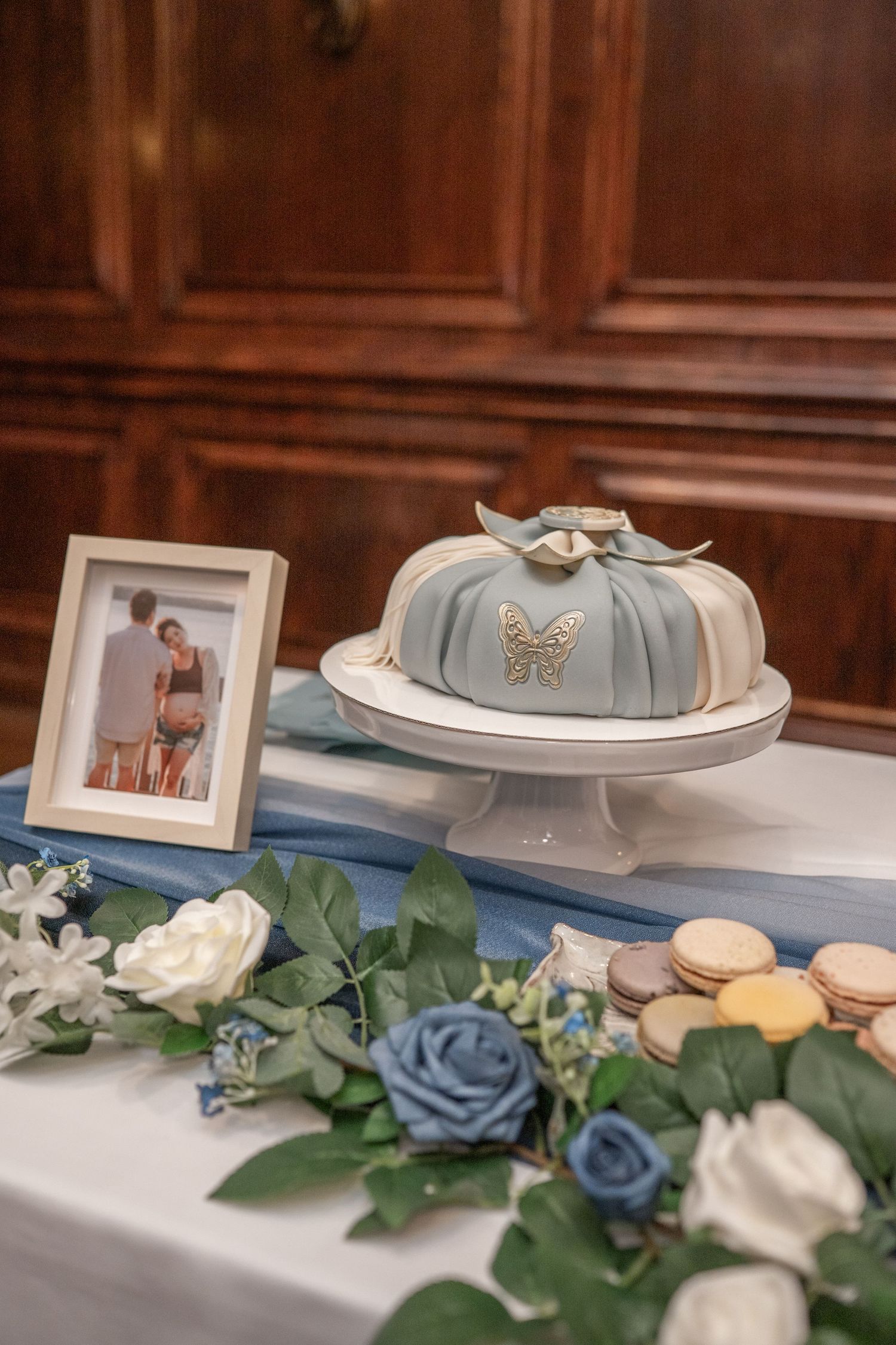 Baby shoes in dusty blue with butterfly applique displayed among white and blue roses and framed photographs. Dohl