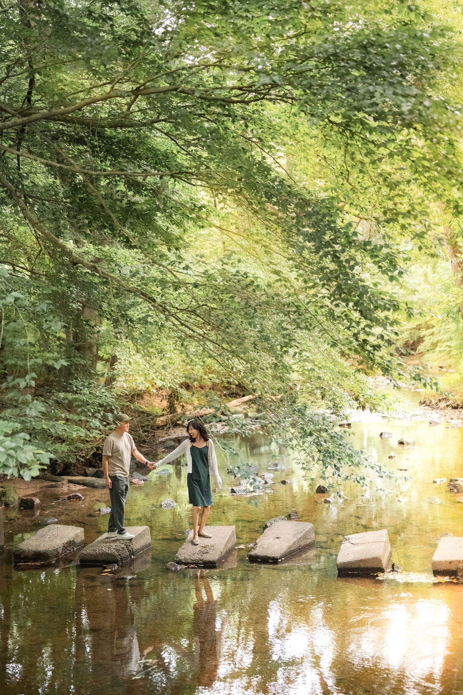 Two people enjoy a tranquil afternoon by the creek, surrounded by lush green trees and water reflections.