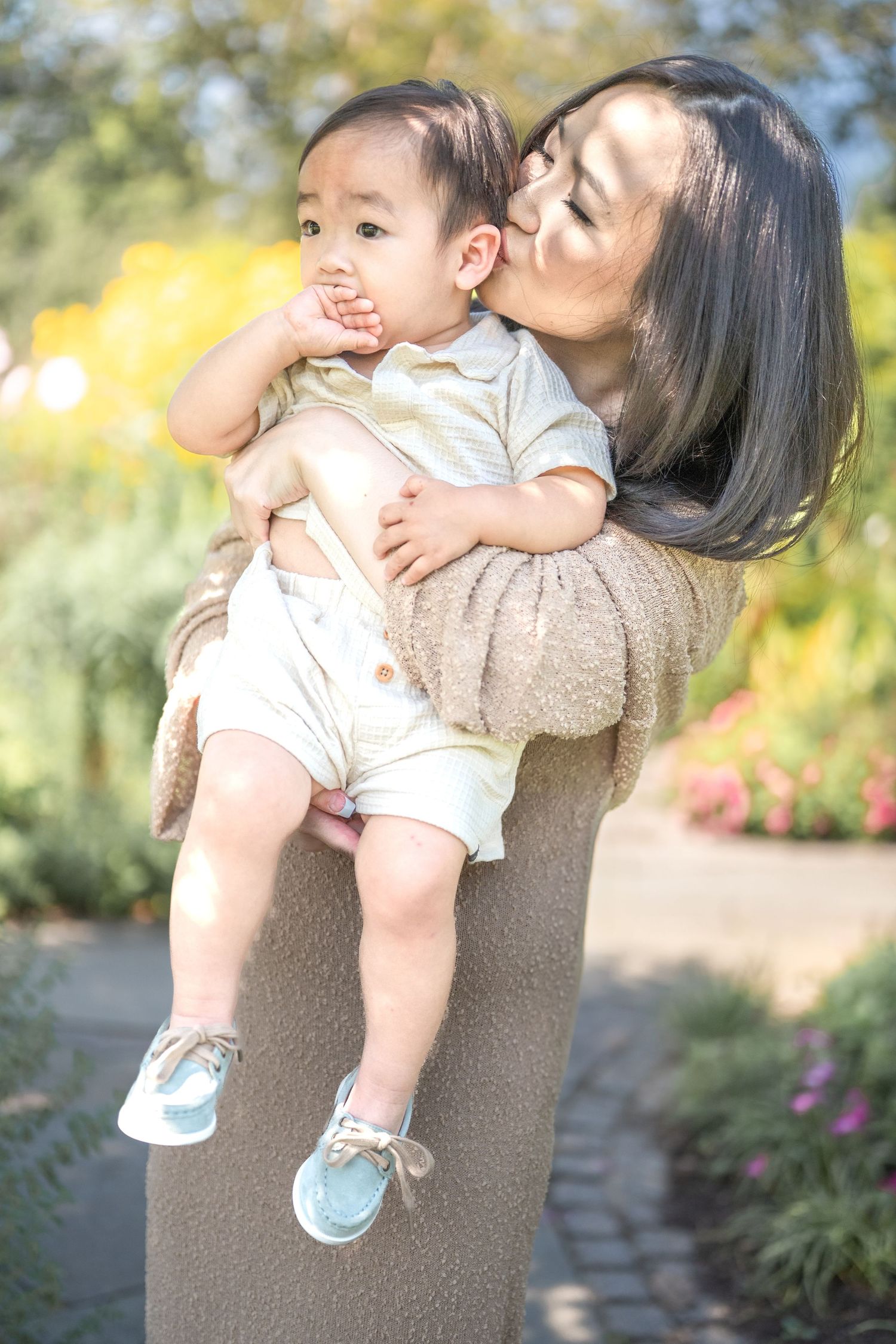 A caregiver lifts and playfully interacts with a toddler in a garden setting with yellow flowers in the background.