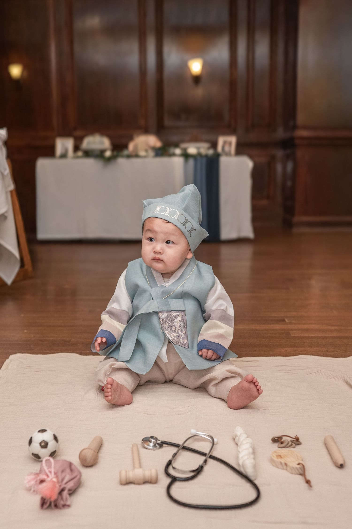 Baby wearing a hat lies on hardwood floor while reaching for various toys and props including a soccer ball and stethoscope. Dohl