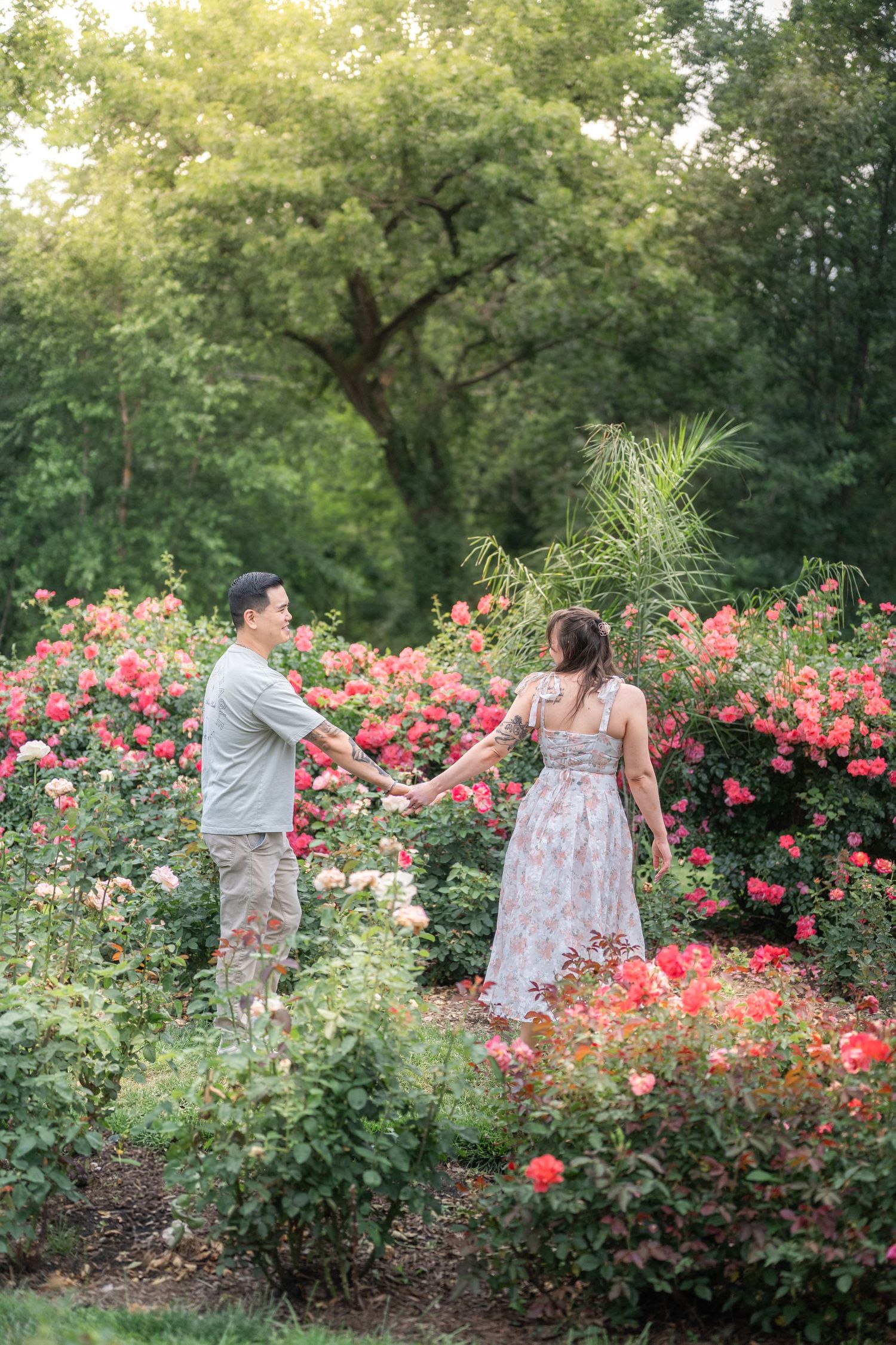Couple shares romantic moment in a lush garden filled with blooming pink roses under leafy green trees.