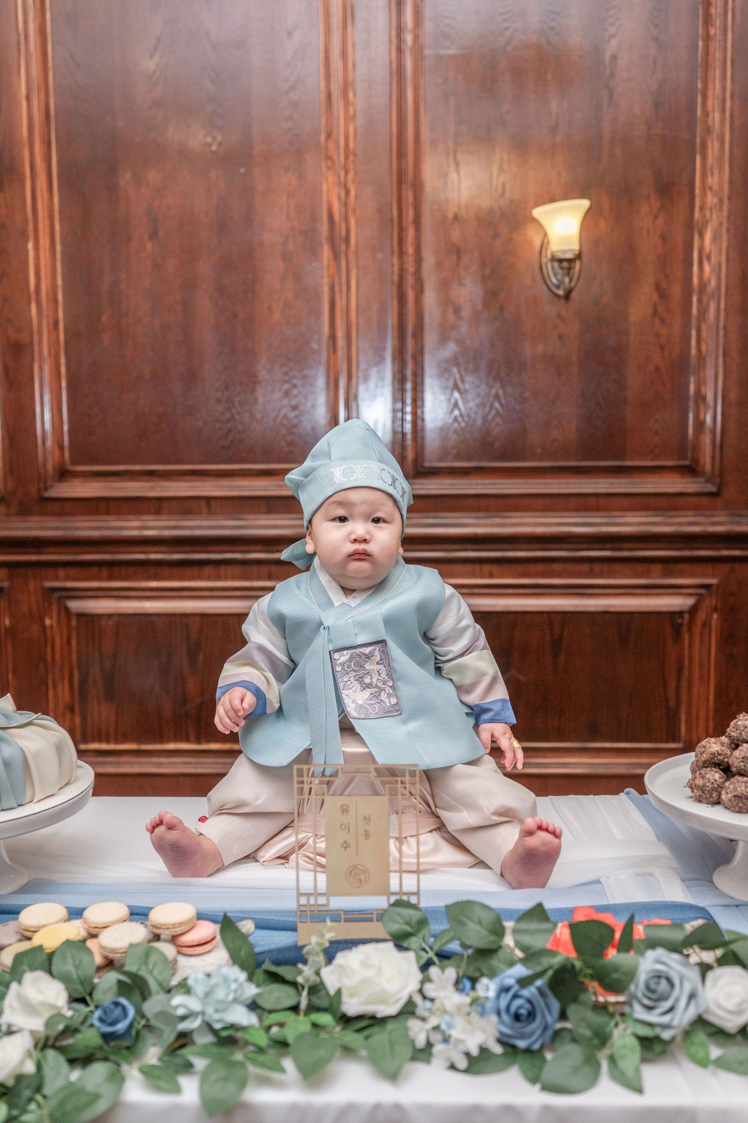 Baby in light blue outfit sits on wooden table decorated with floral garland at formal event setting.