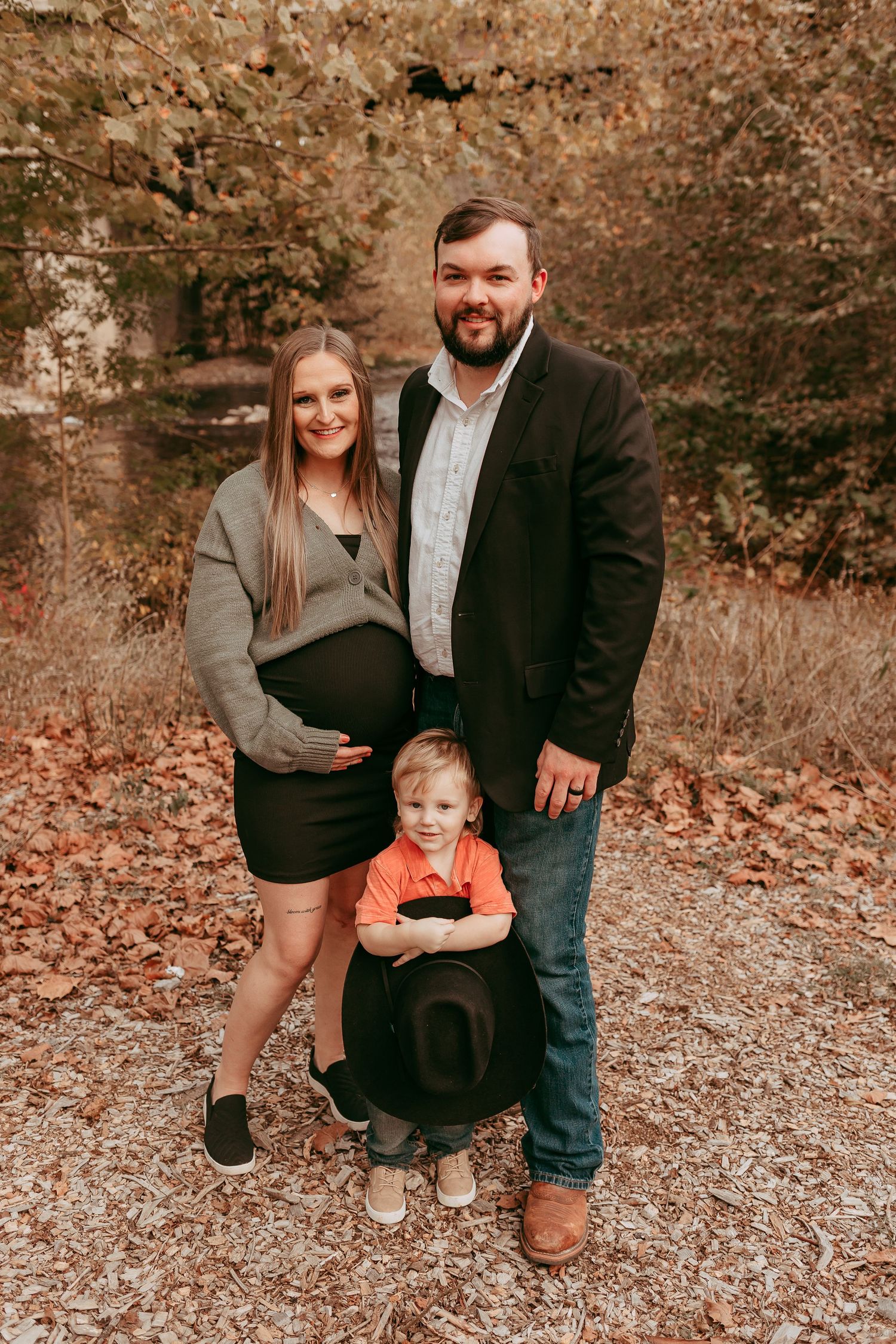 Pregnant woman, bearded man, and young child pose together outdoors among autumn foliage.