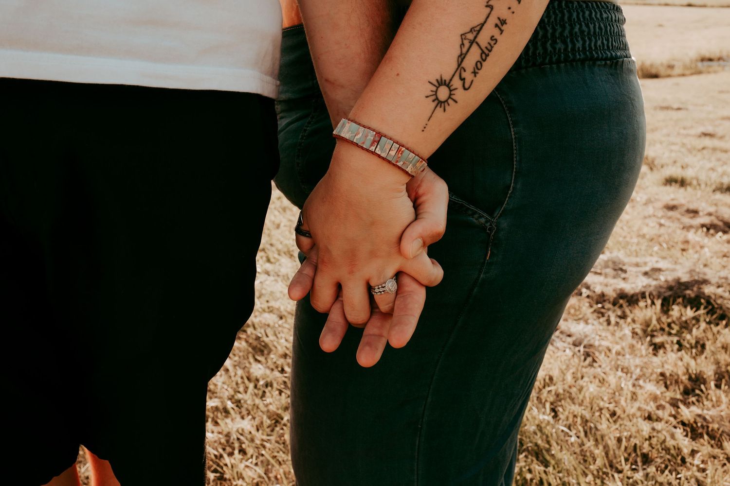 Close-up of couple holding hands showing tattoos, bracelet, and engagement ring in outdoor field setting.