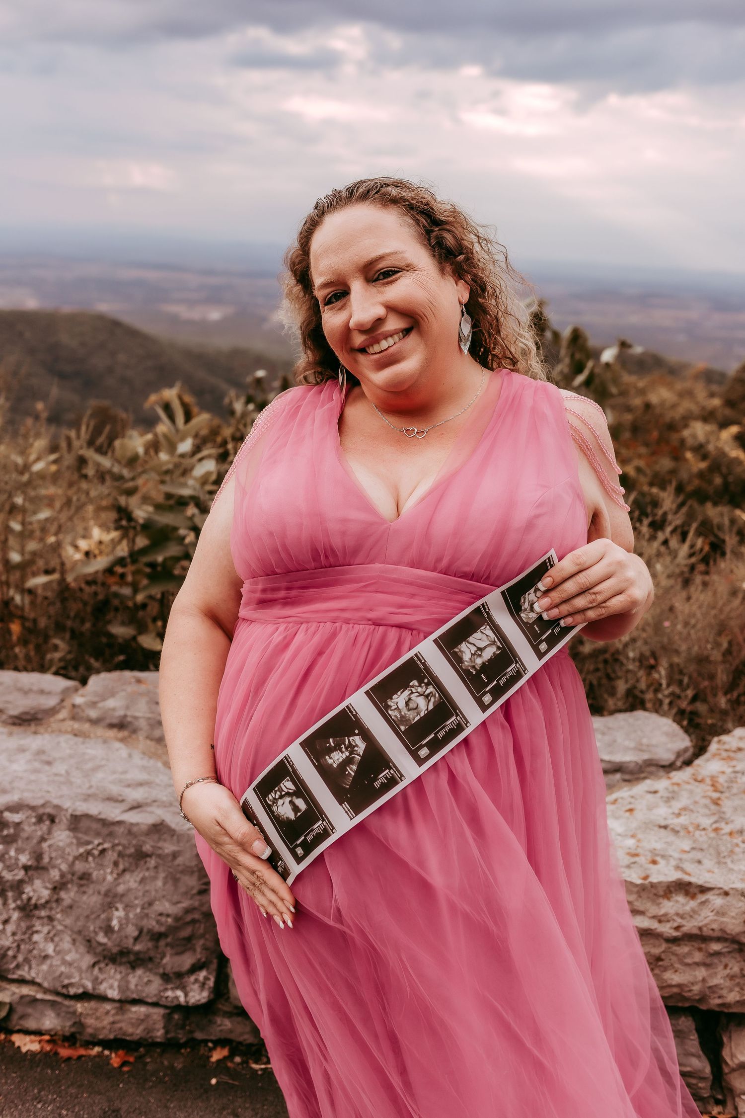 Smiling pregnant woman in pink dress holding ultrasound photos at scenic mountain overlook.