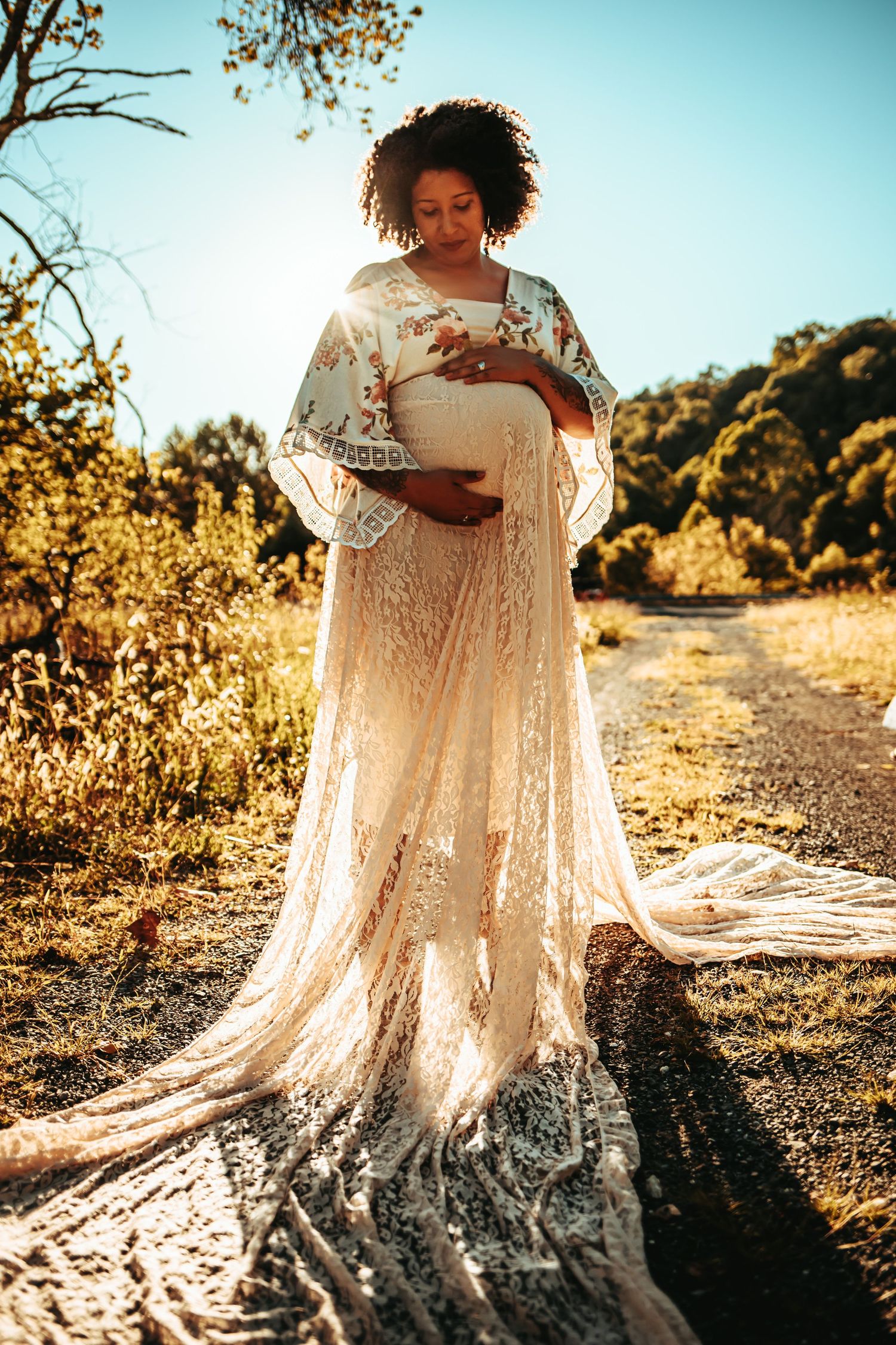 Backlit pregnant woman in flowing lace dress holding belly on sunny outdoor trail.