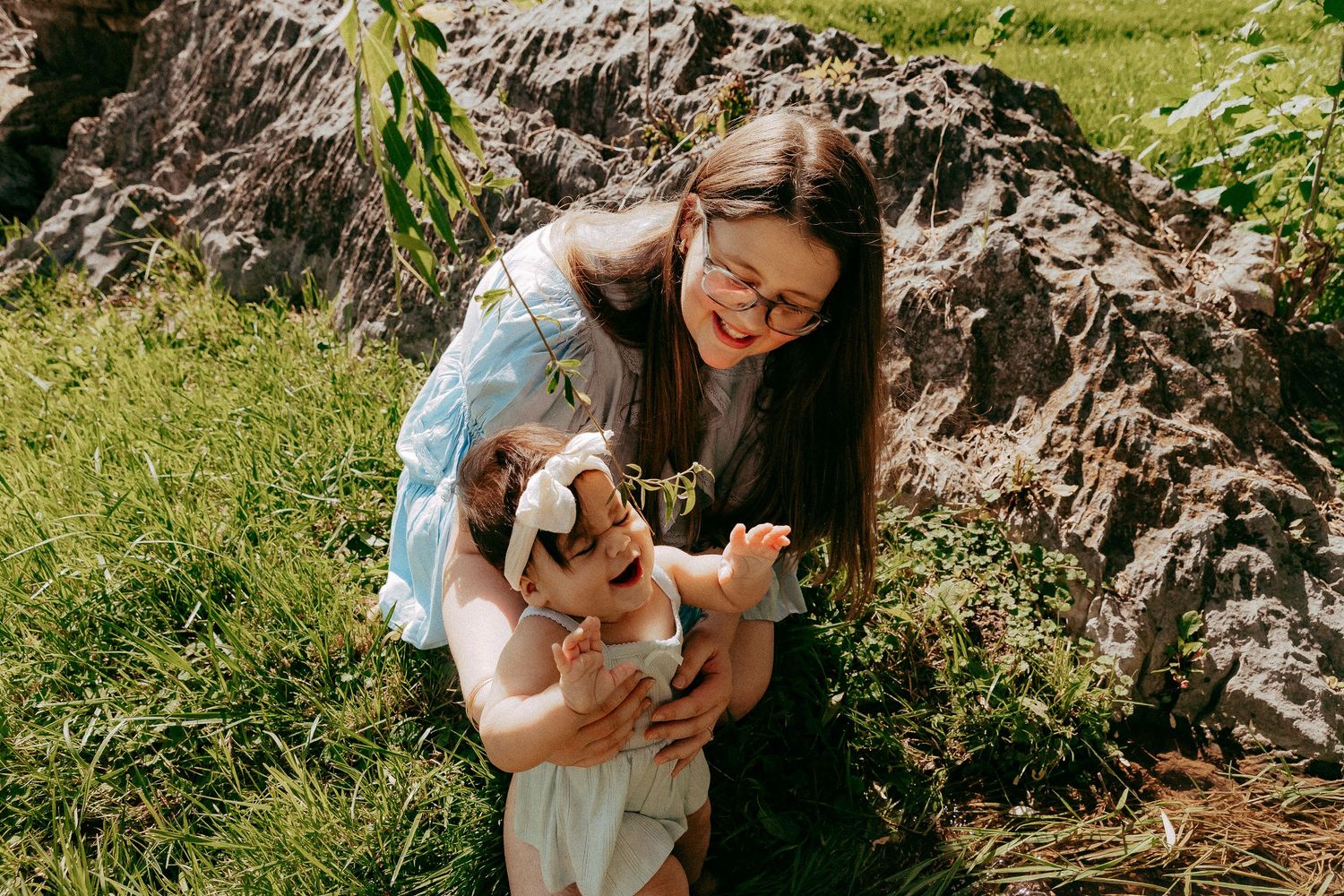 A smiling woman with glasses holds a laughing baby outdoors near rocky terrain and green grass on a sunny day.