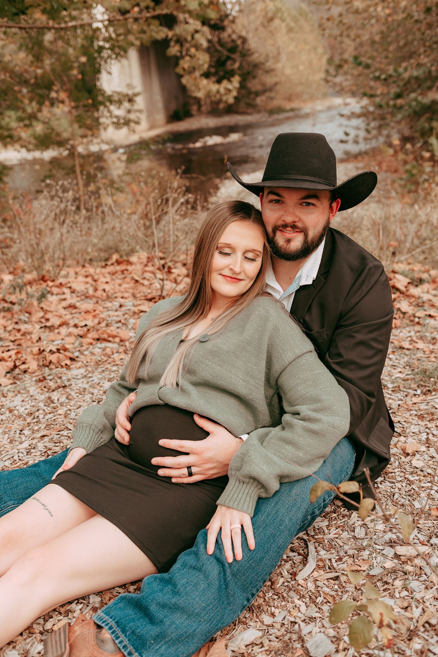 Pregnant couple sitting together in autumn leaves, man in cowboy hat embracing woman.