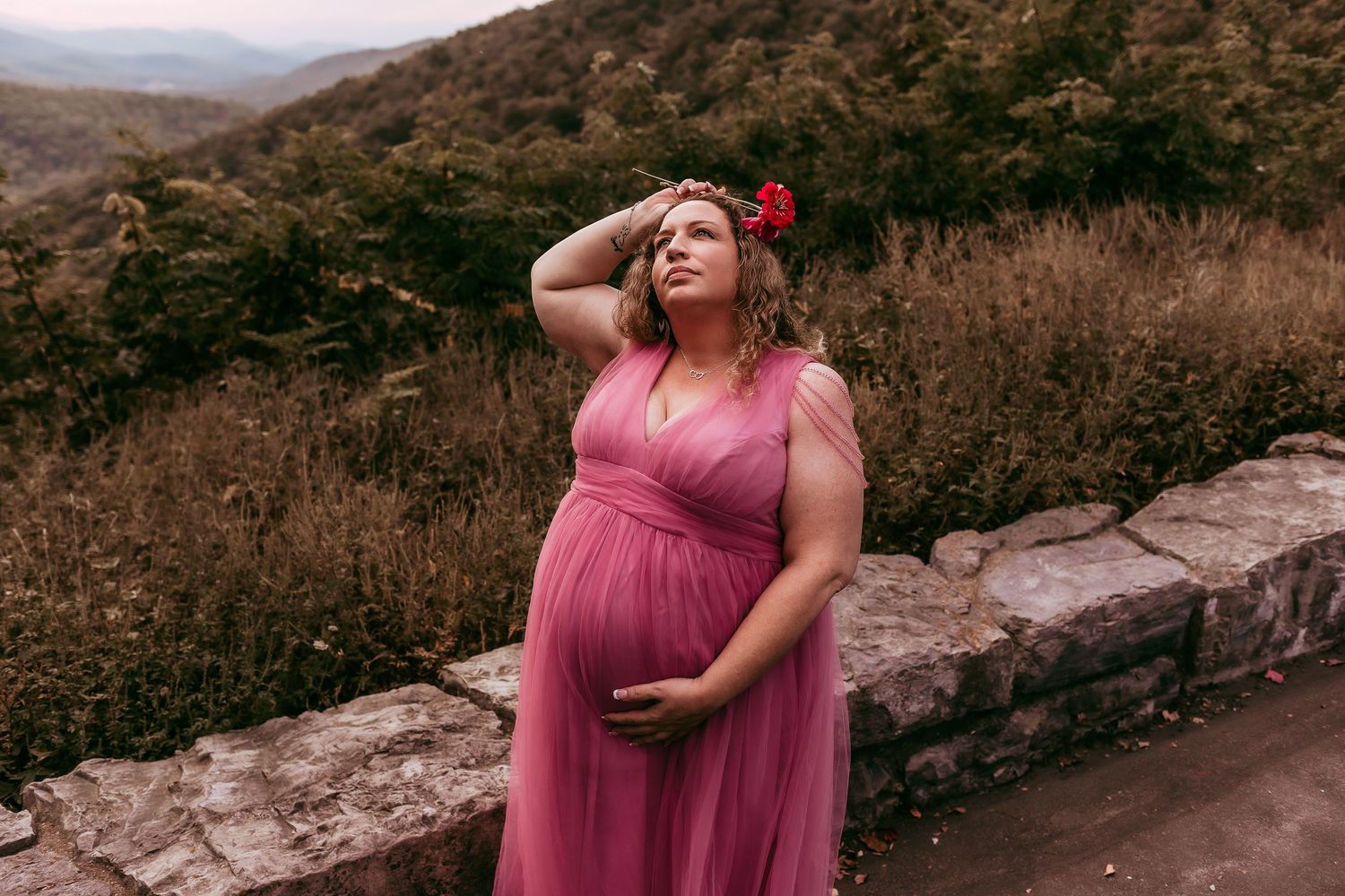 Pregnant woman in pink tulle dress with red flower crown at mountain overlook setting.