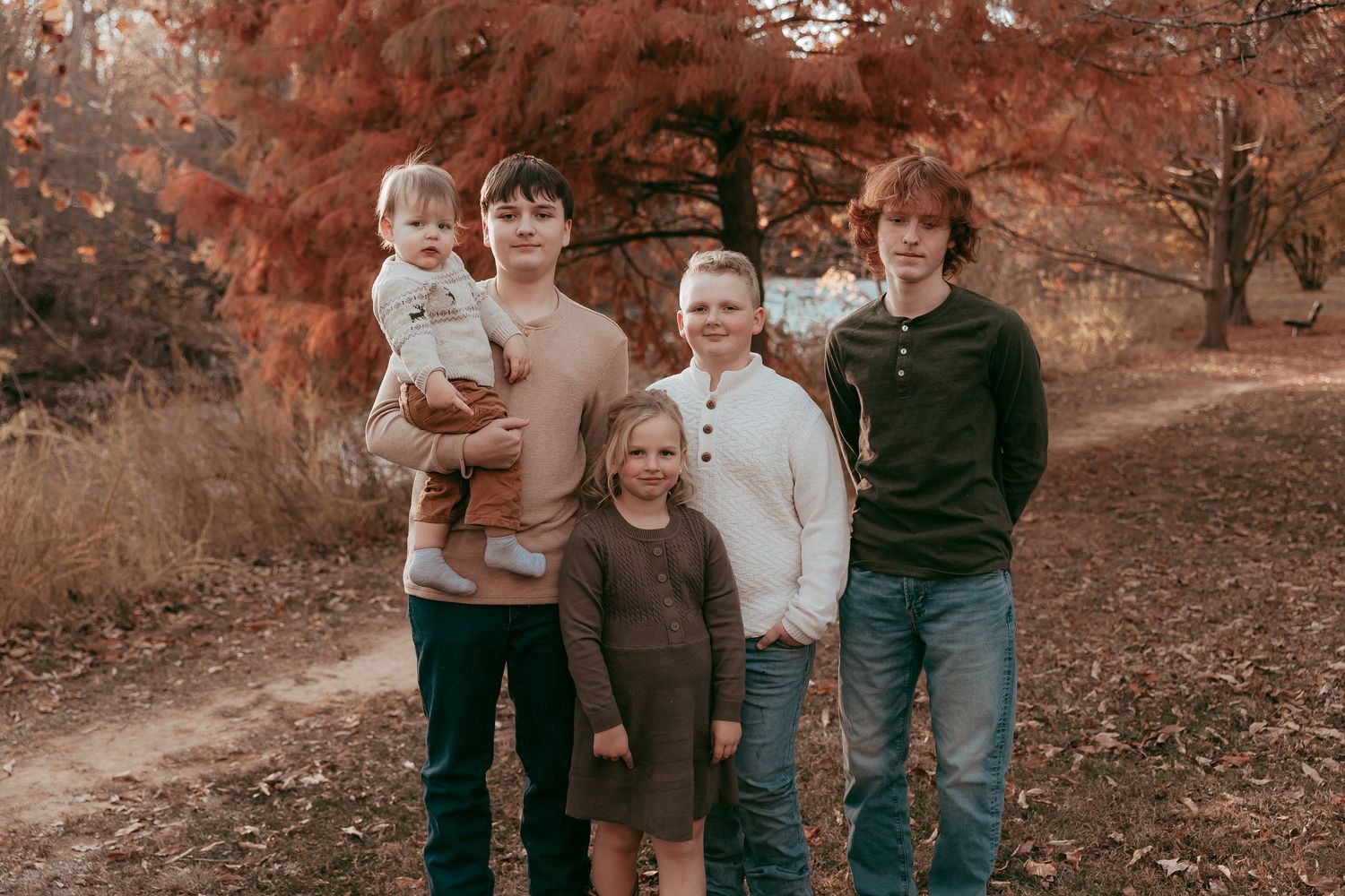 Five siblings pose together on a path surrounded by warm red and orange fall trees.
