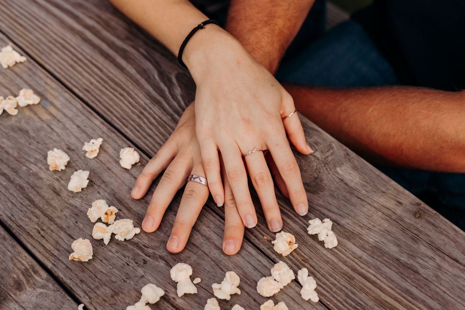 Engaged couple showing off diamond ring with hands placed on rustic wooden table with popcorn.