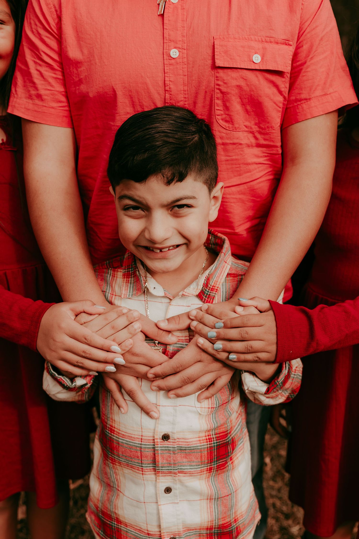 Smiling boy in plaid shirt being hugged by family members in red outfits.