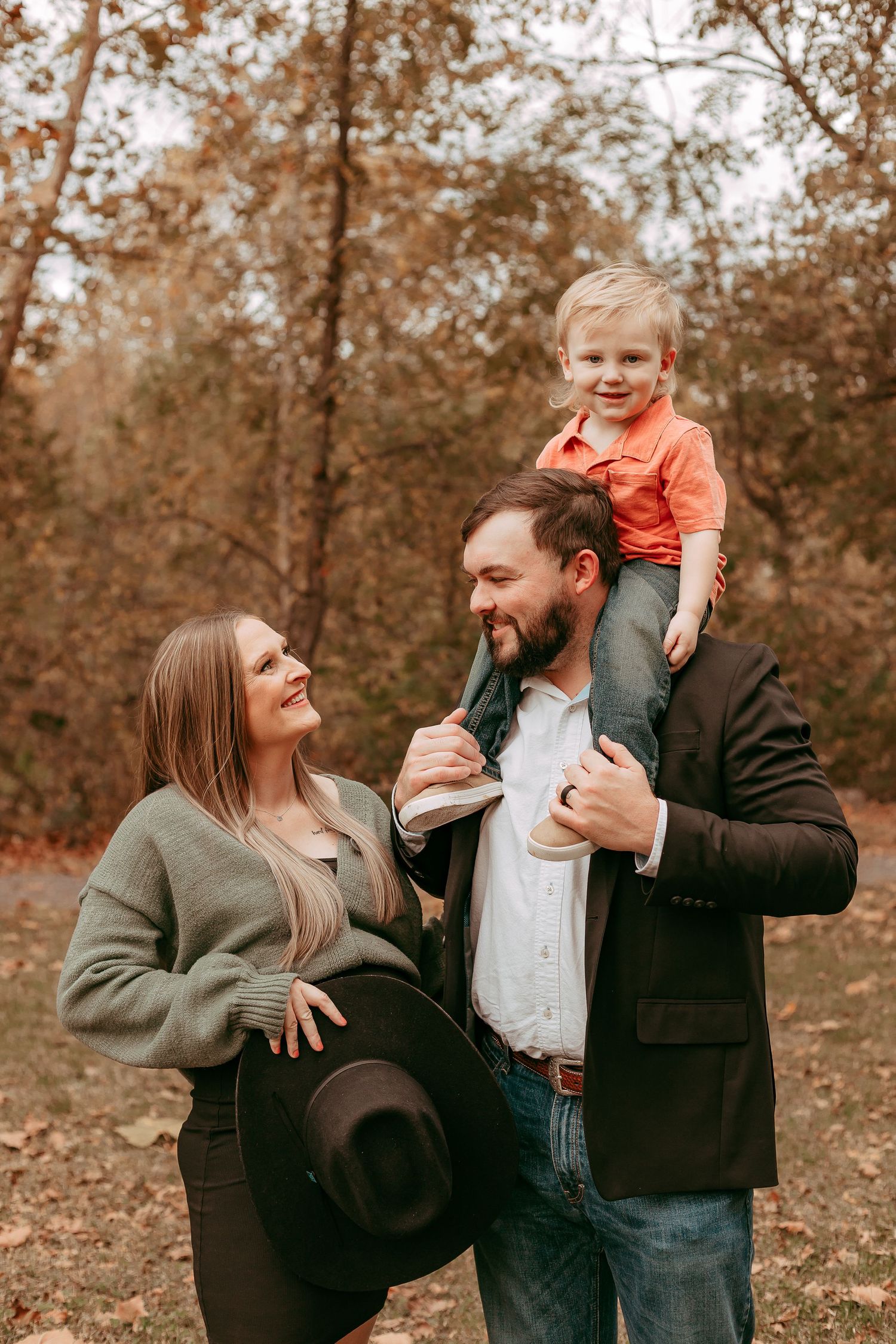 Happy family of three in autumn forest, father carrying toddler son on shoulders beside smiling mother.