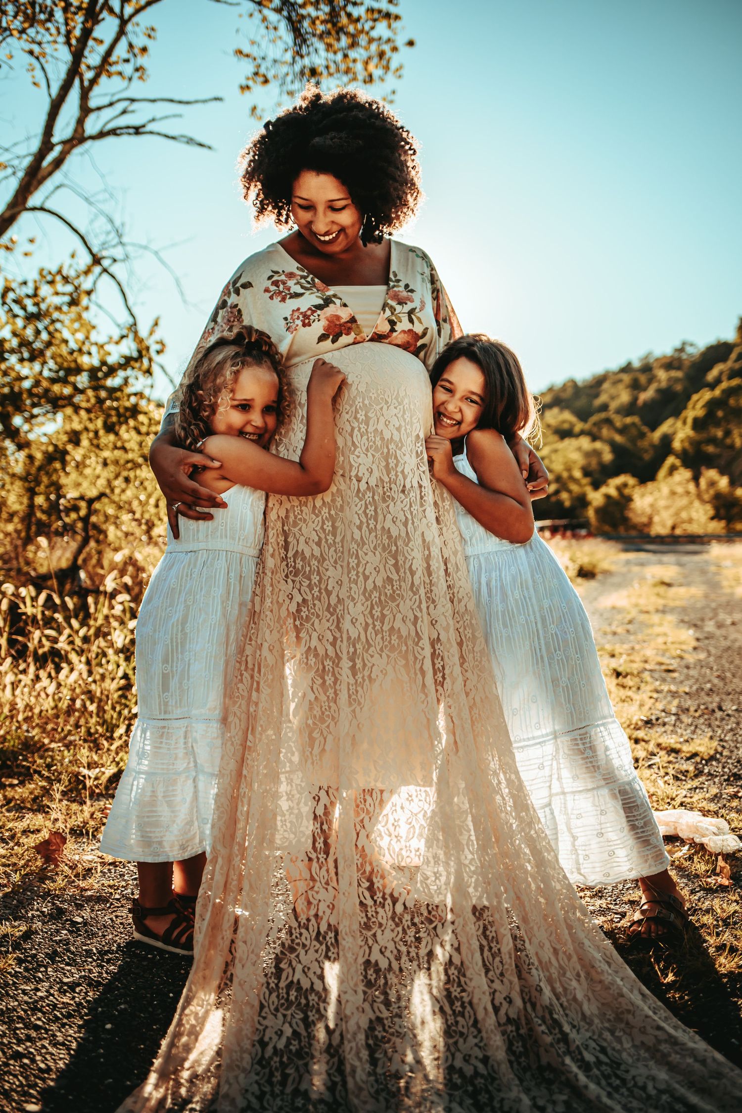 Smiling pregnant woman in white lace dress embraced by two young girls in matching white dresses outdoors.