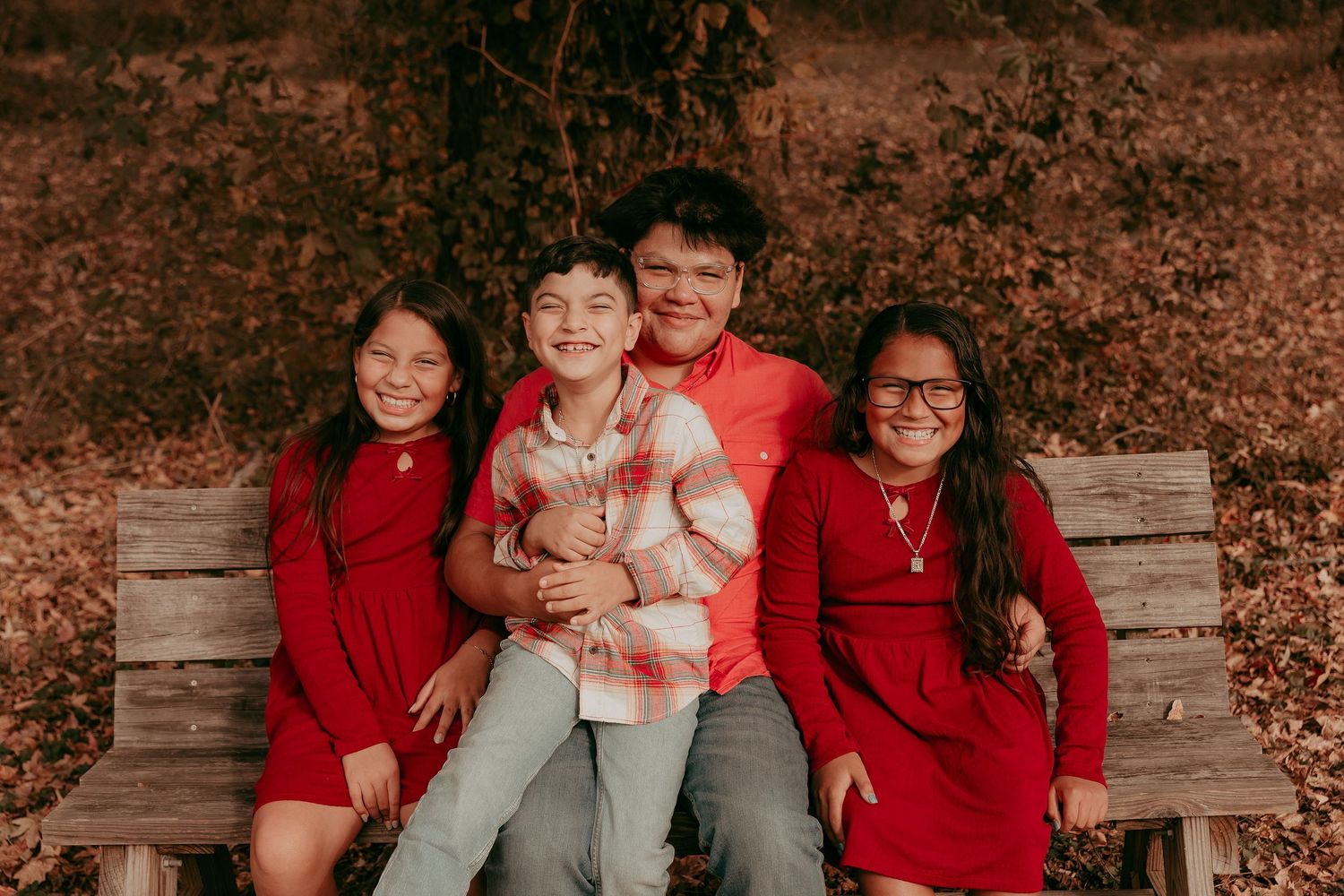 Four siblings posing together on a rustic wooden bench surrounded by fall foliage.
