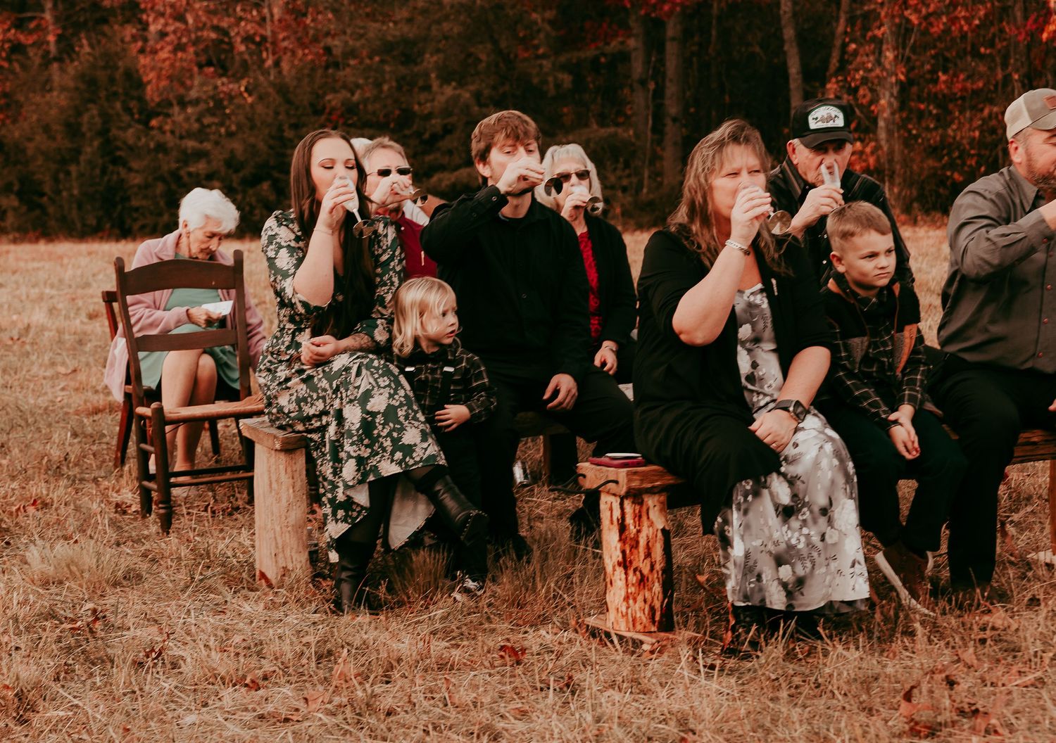 Wedding guests seated on wooden benches blowing bubbles during an intimate outdoor autumn ceremony surrounded by fall foliage.