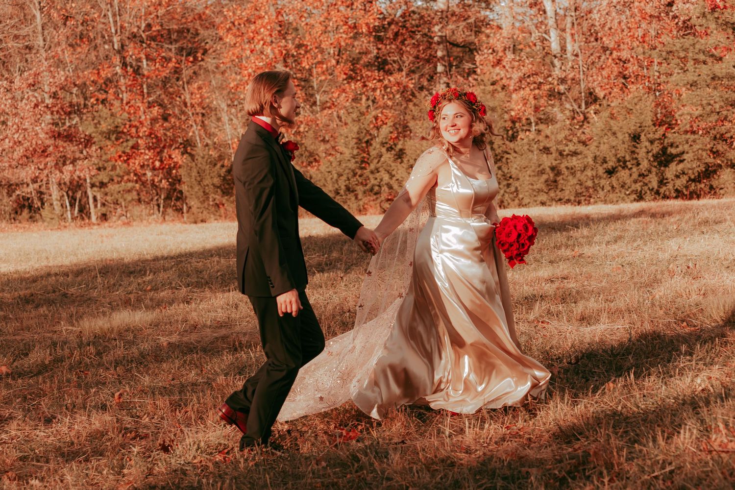 Bride in champagne gown and groom in black suit holding hands running through autumn field with red foliage backdrop.