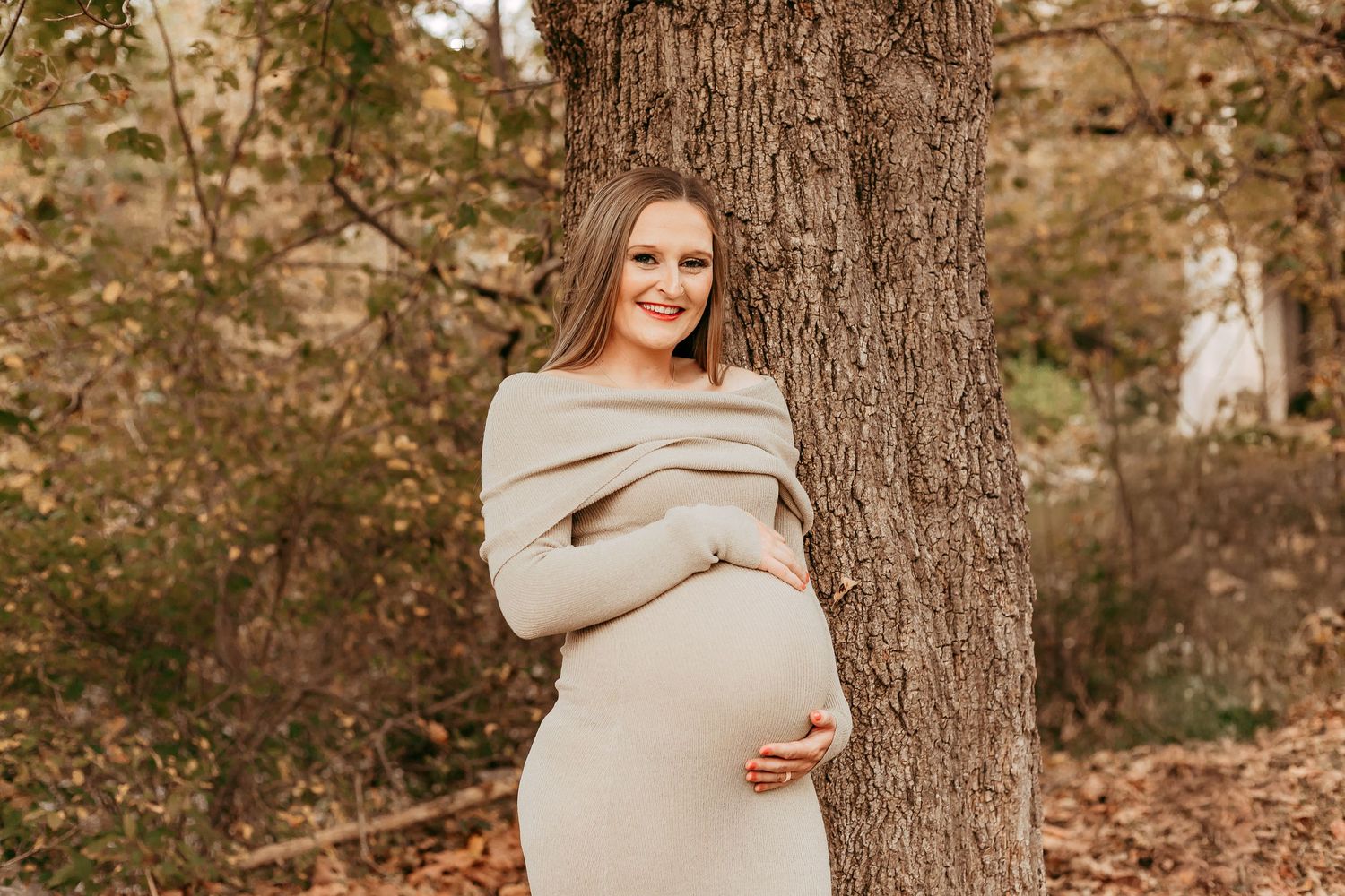 Smiling pregnant woman in beige off-shoulder dress leaning against tree in fall setting.