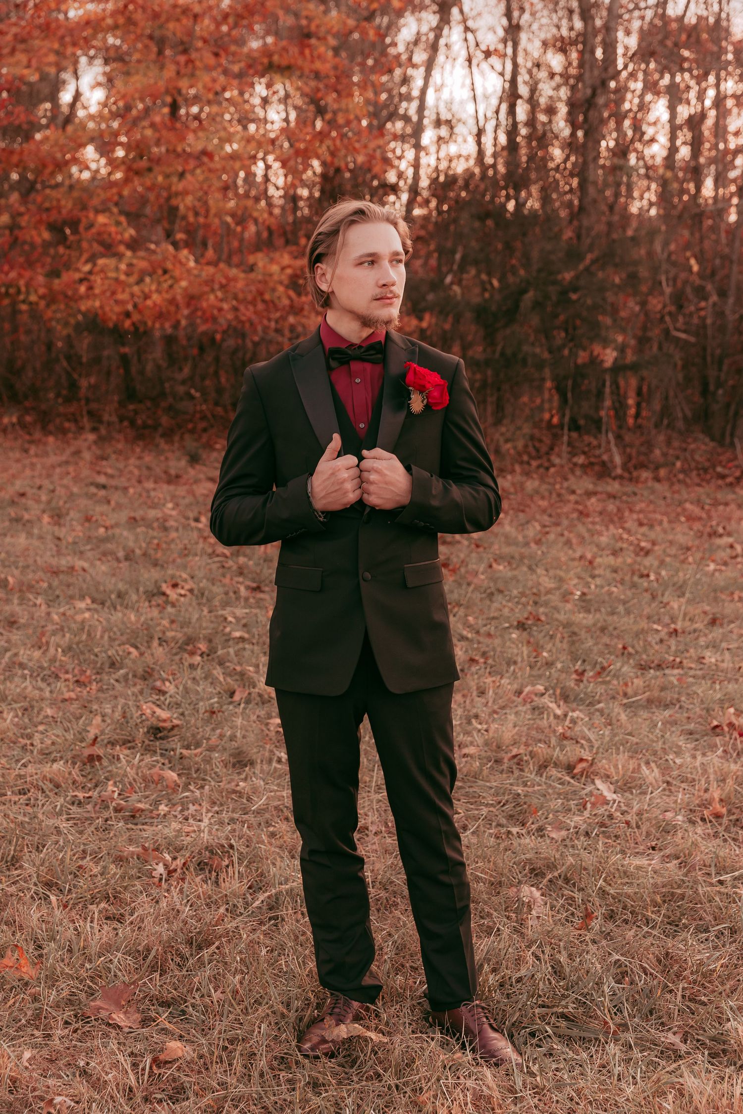 Groom in black tuxedo adjusts jacket lapels standing in autumn outdoor setting.