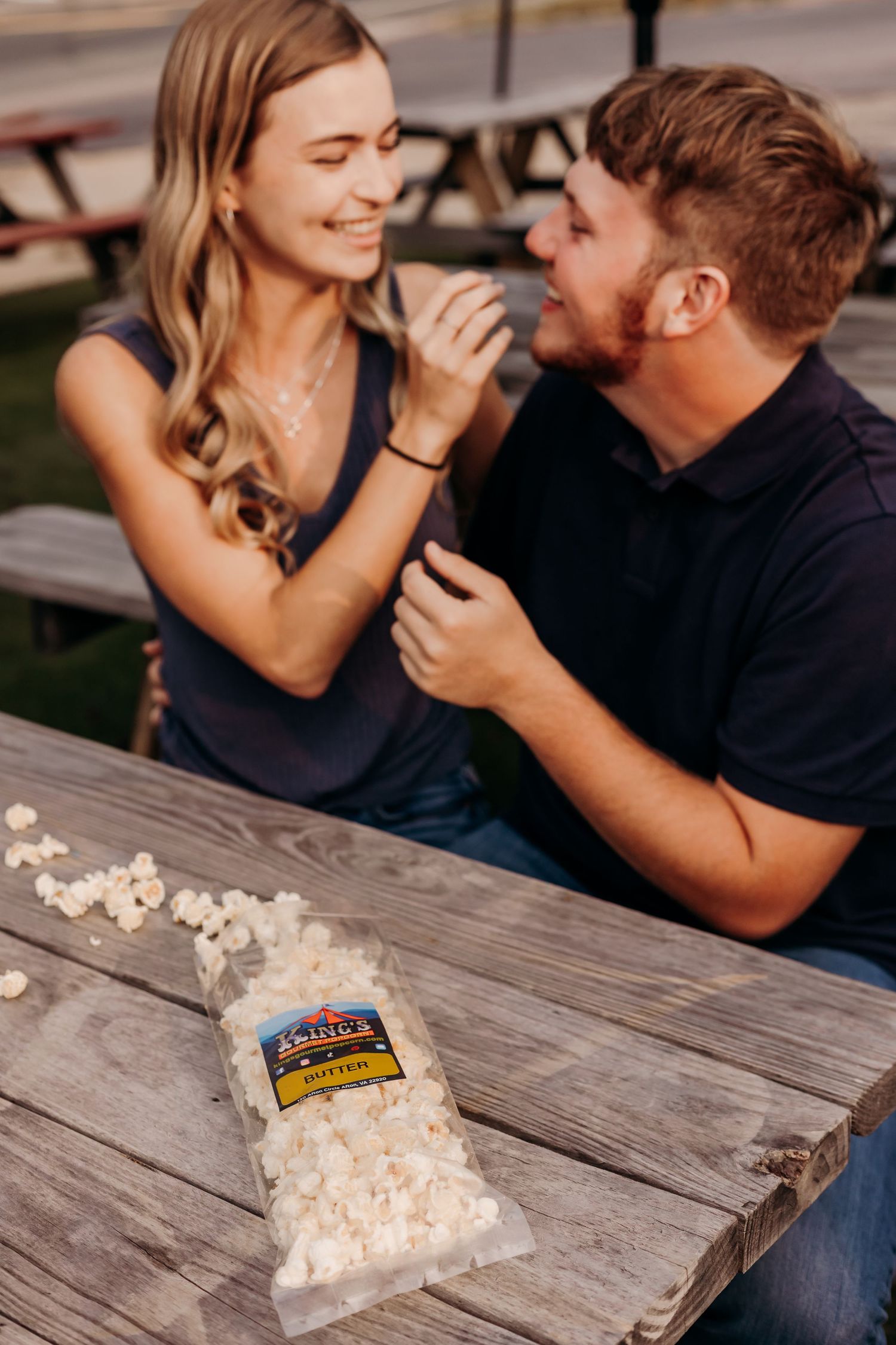 Couple sharing popcorn at a picnic table, smiling and enjoying a romantic outdoor date.