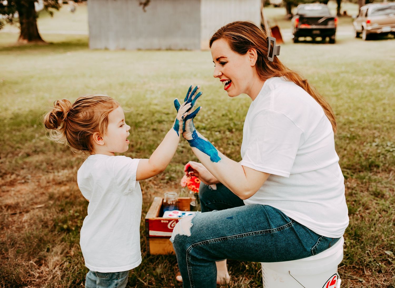 A joyful pregnant woman and her paint-covered toddler share a high-five outdoors in a grassy yard.