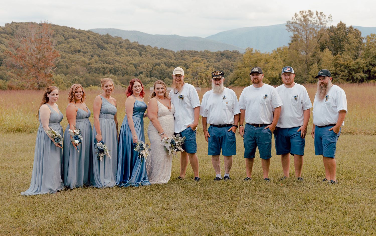 Wedding party poses together in a mountain field, bridesmaids in blue gowns and groomsmen in white shirts.