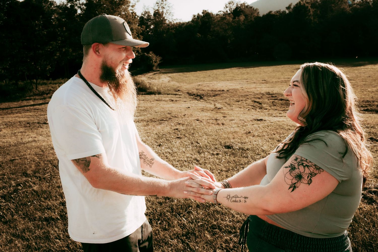 Tattooed couple holding hands and smiling at each other in sun-drenched field with mountain backdrop.