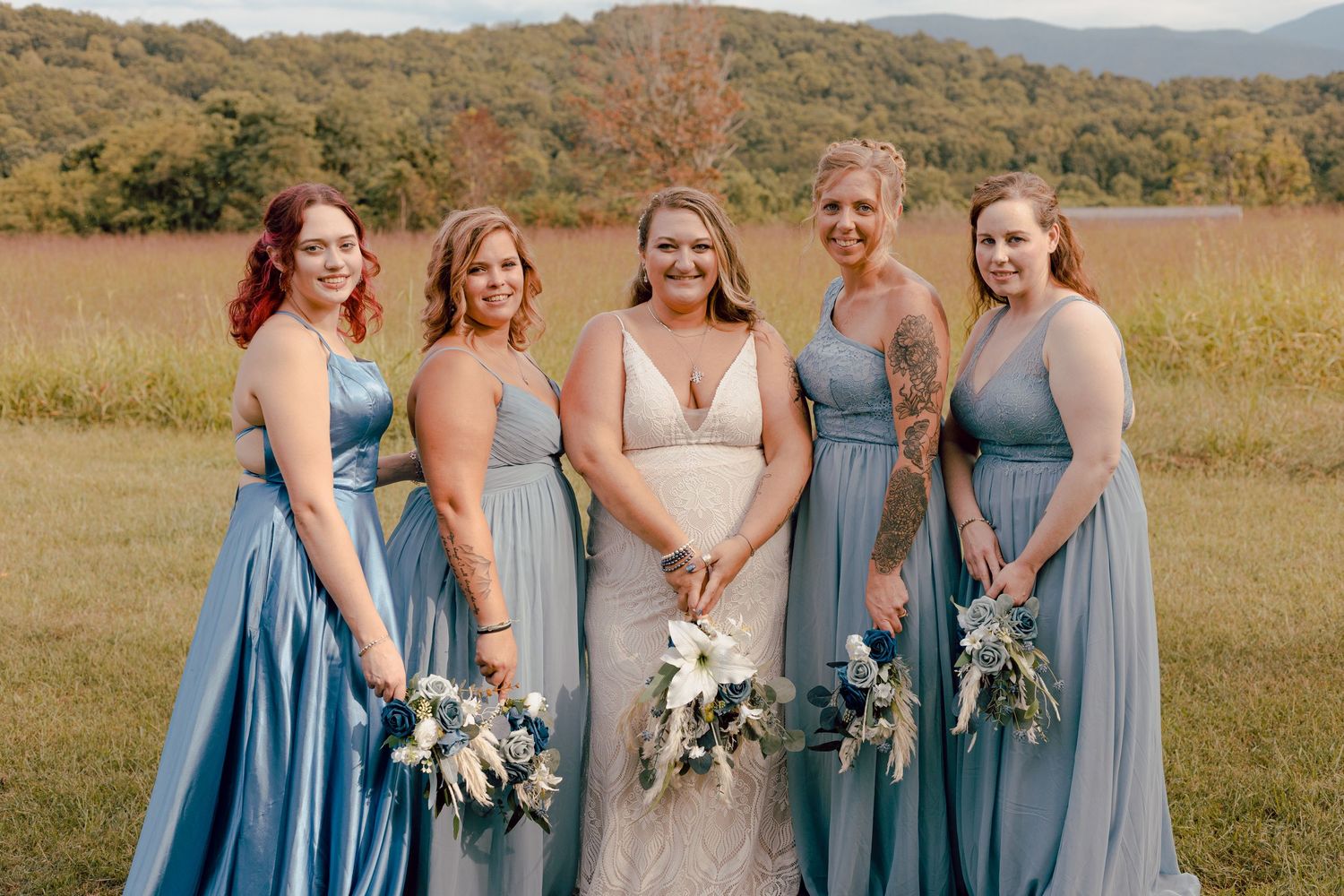 Bride poses with four bridesmaids in dusty blue gowns in a scenic mountain field wedding setting.