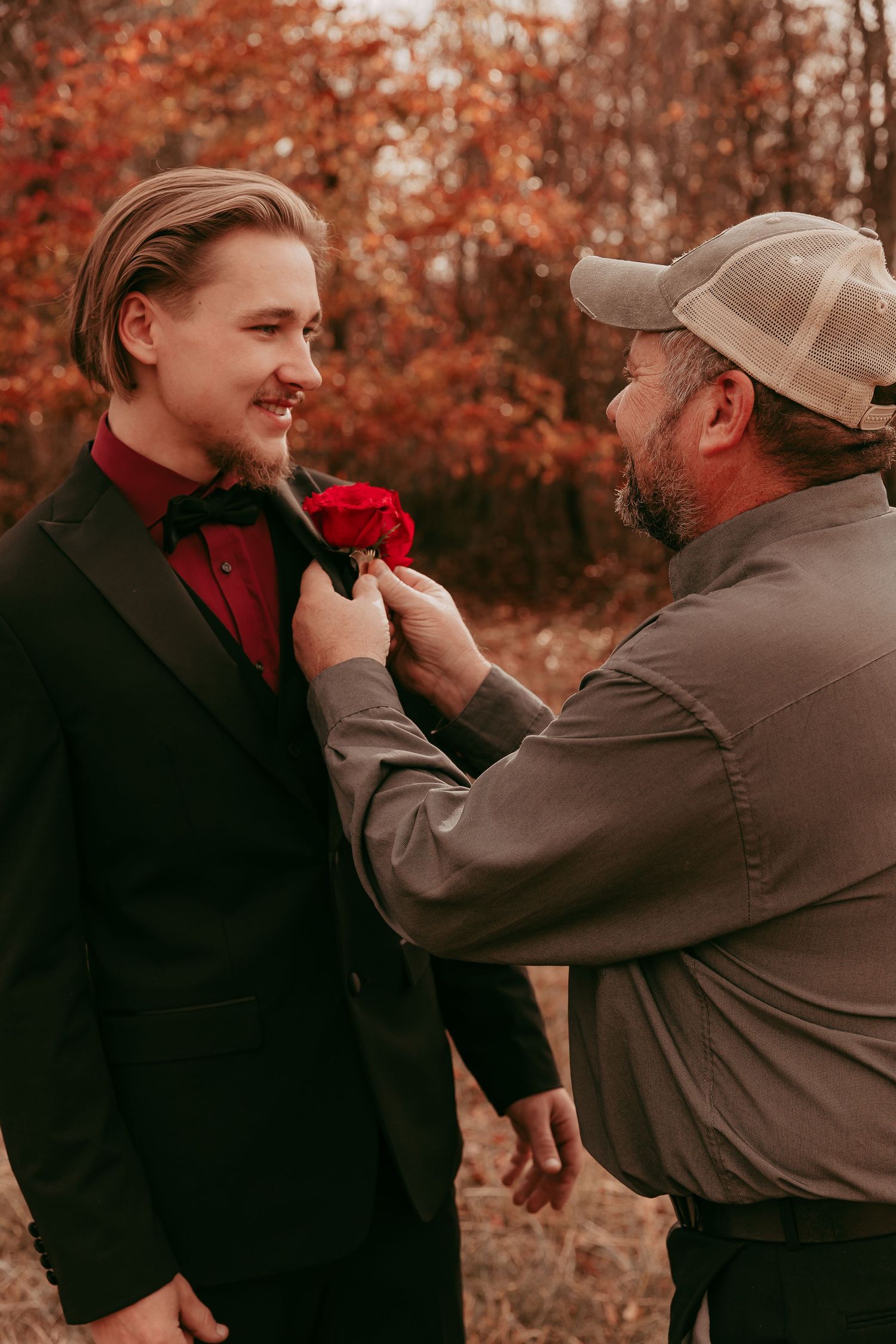 Father pins red rose boutonniere on groom's black tuxedo before fall wedding.