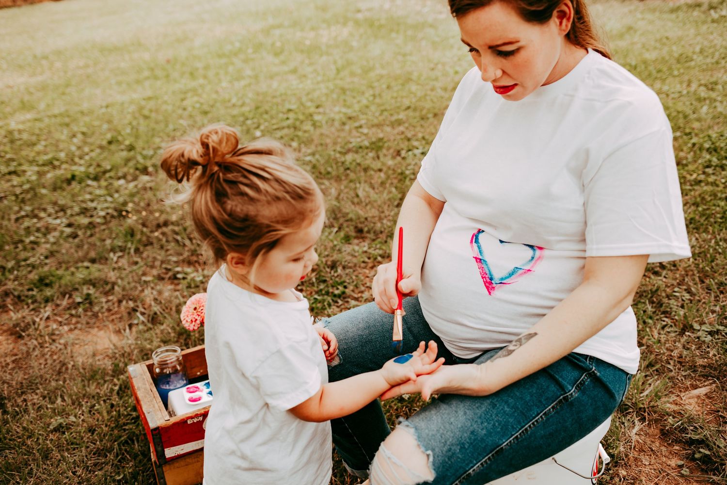 A pregnant woman in a white t-shirt lets her toddler paint a colorful heart on her baby bump outdoors.