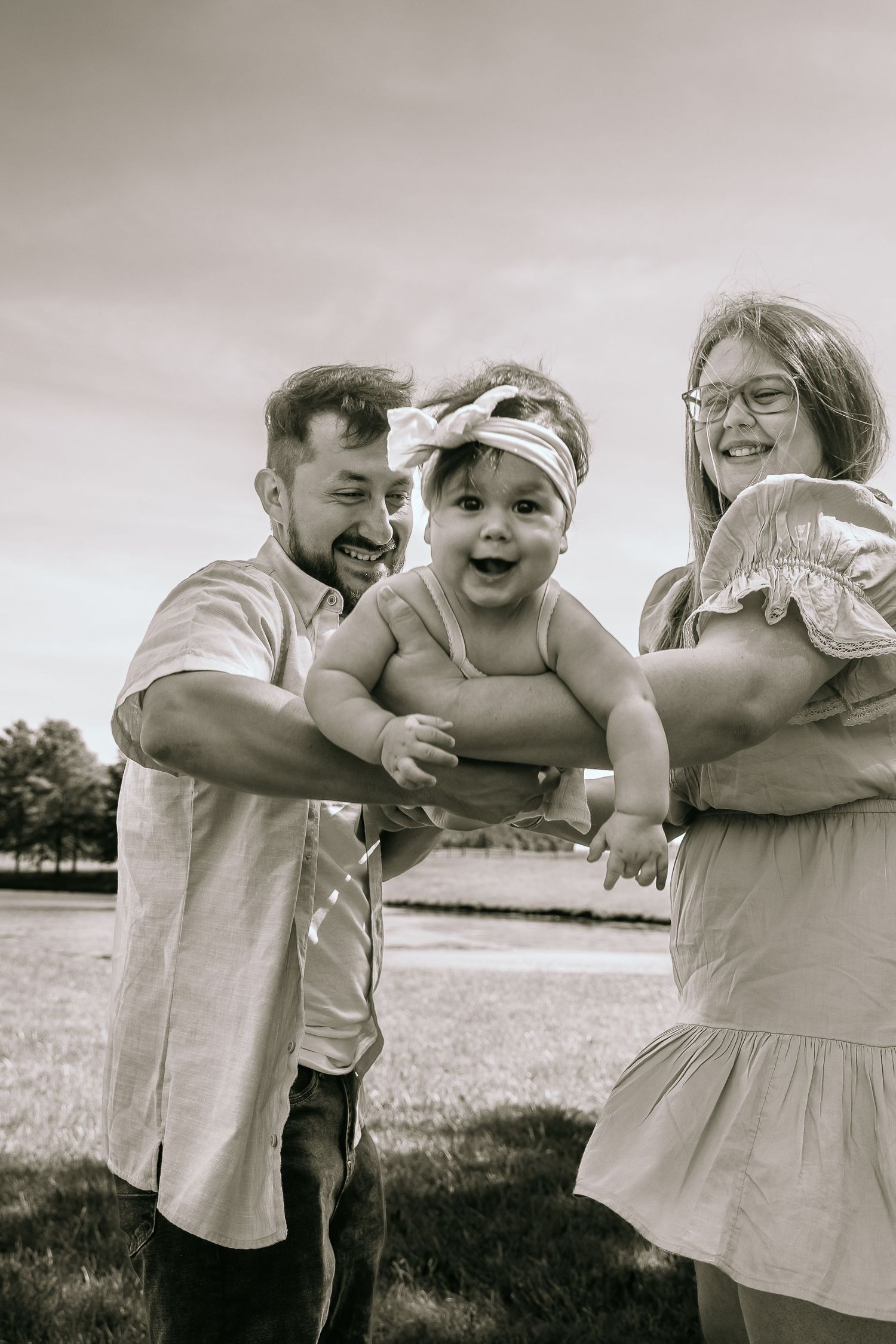 Black and white photo of happy parents holding their laughing baby girl outdoors in a park setting.
