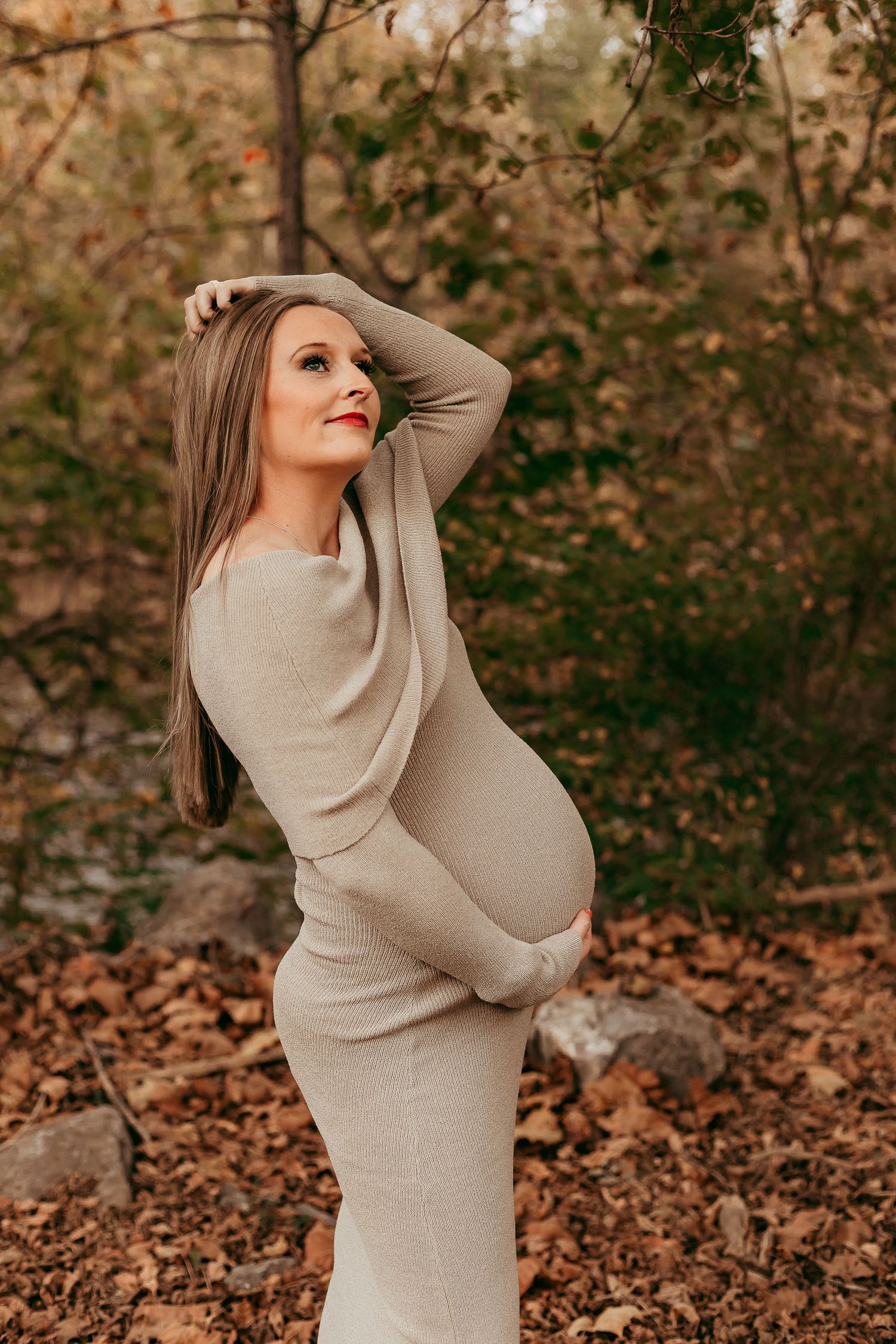 Pregnant woman in beige ribbed dress cradling baby bump among colorful autumn foliage outdoors.