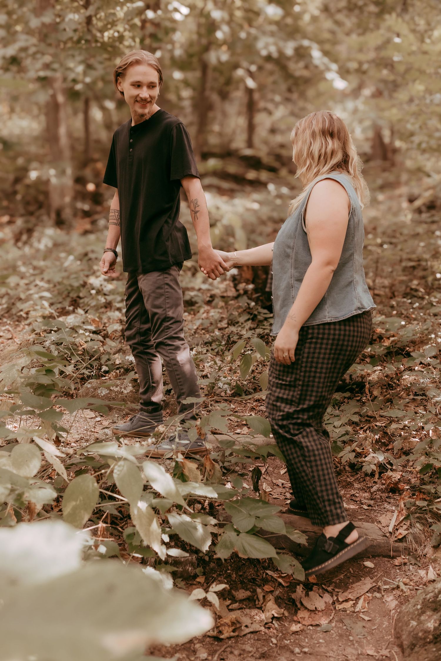 Couple holding hands and walking through lush green forest trail smiling together.