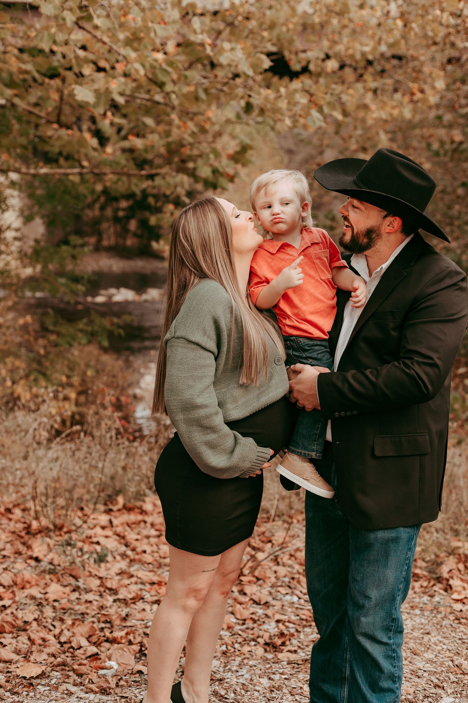 Couple kissing their blonde toddler son surrounded by colorful autumn leaves outdoors.