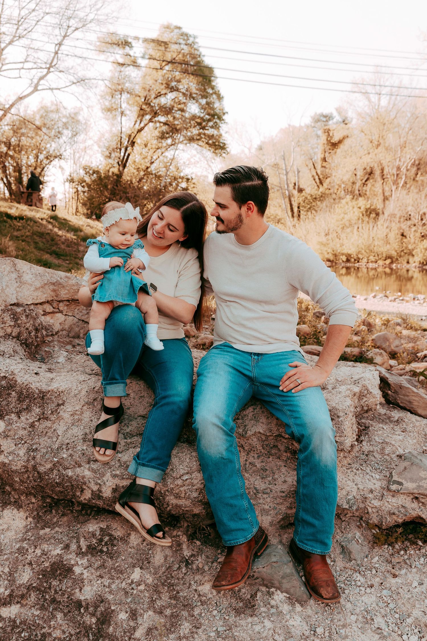 A young family with a baby girl sits on rocks outdoors, smiling warmly in casual jeans and light tops.