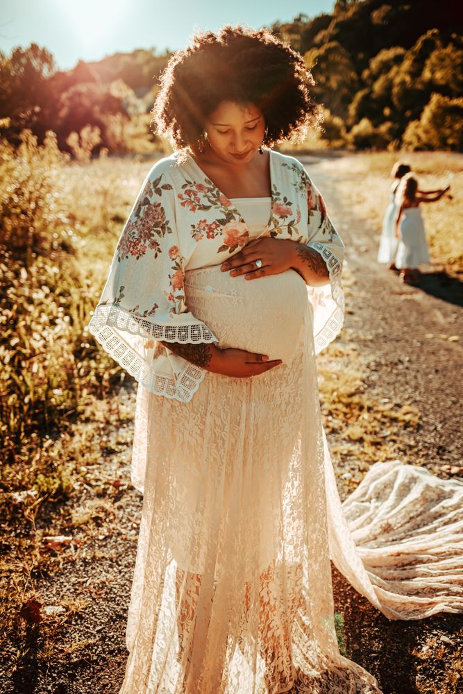 Pregnant woman in lace maternity dress and floral wrap cradling belly on sunlit dirt path.